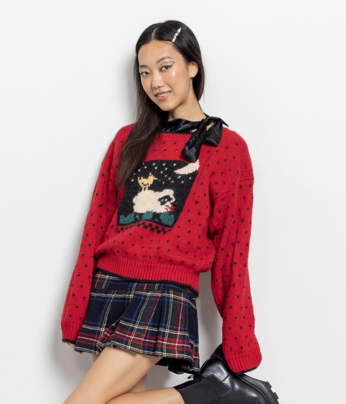Person wearing a red sweater with a graphic design, black skirt, and black boots on a white background