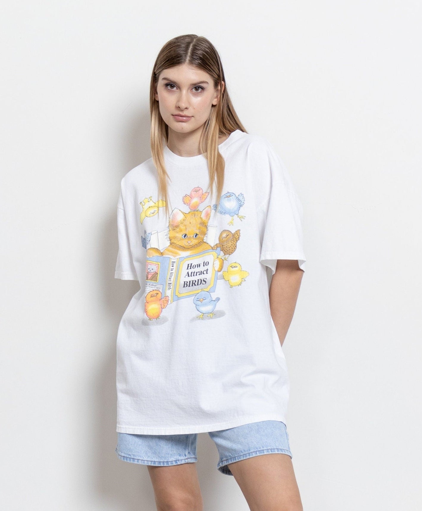 A woman modeling a vintage white t-shirt with a graphic that says 'How to Attract Birds' featuring cartoonish cat characters.