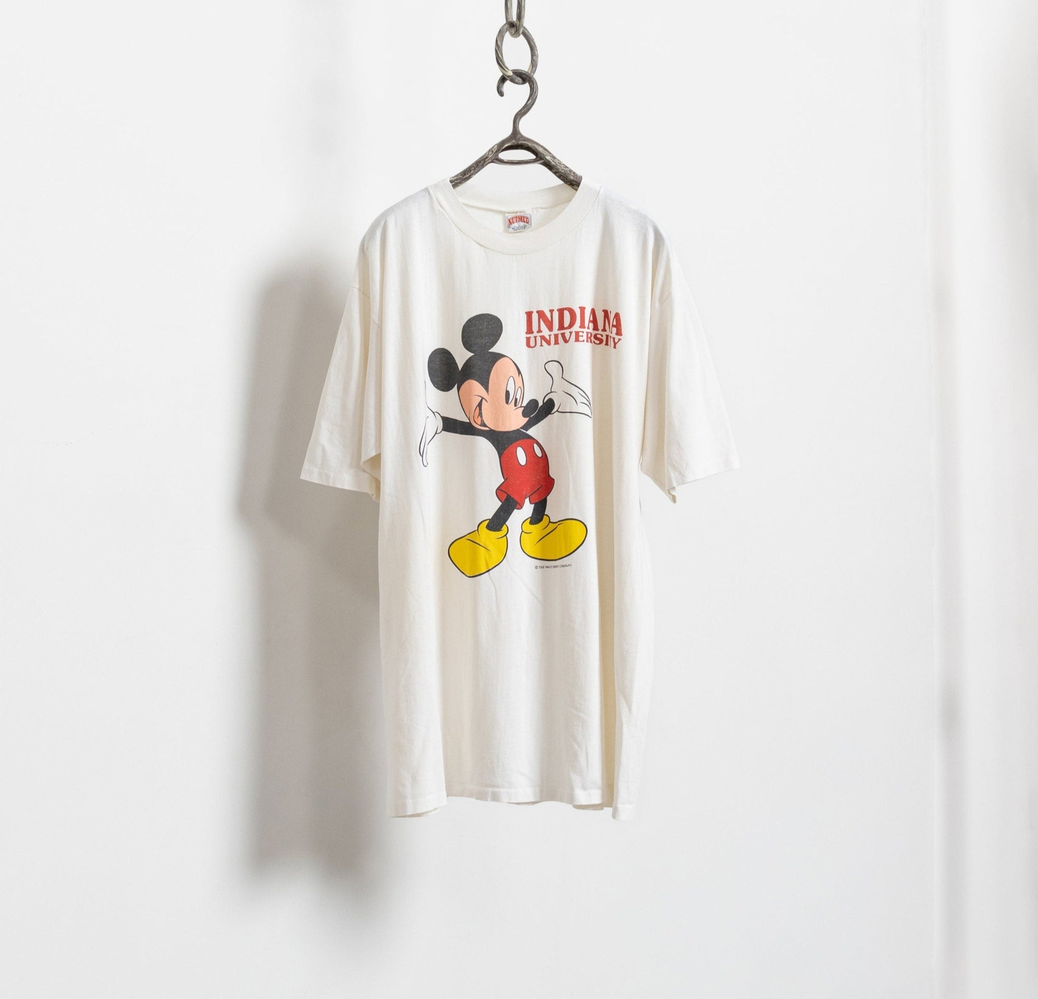 A white vintage Mickey Mouse Indiana University graphic tee hanging on a wall, featuring a graphic of Mickey Mouse in red and yellow shorts with the text 'Indianapolis'.