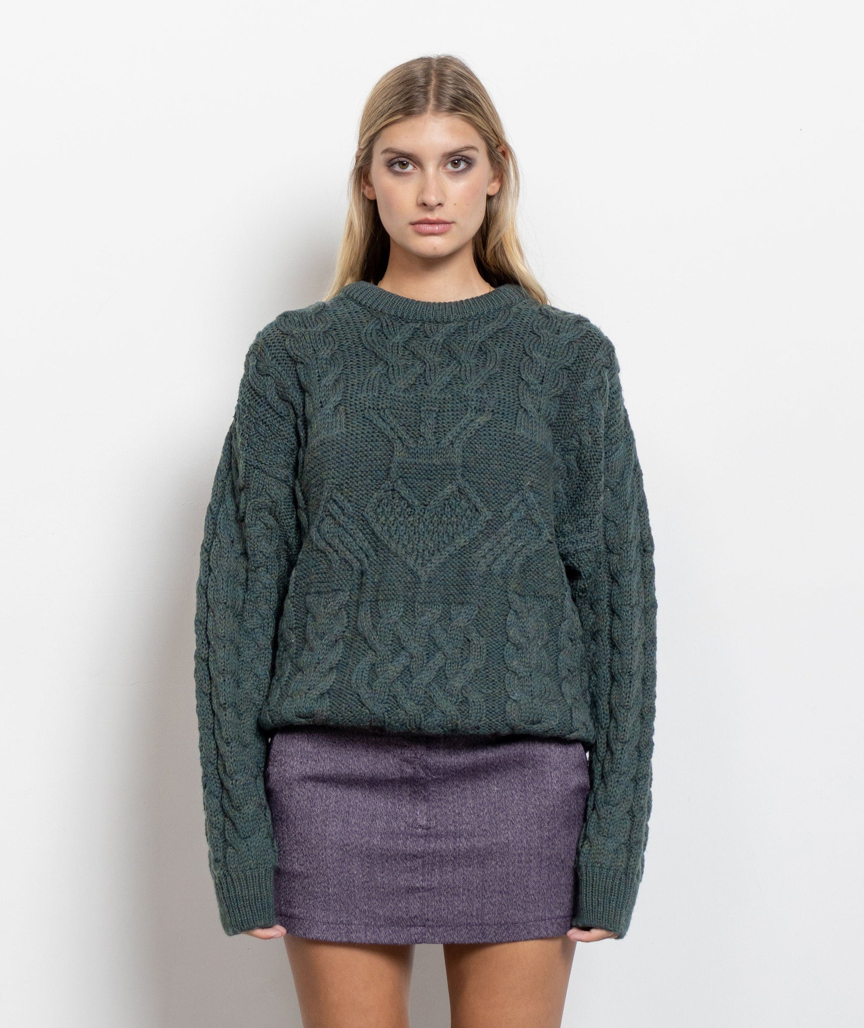 Woman wearing a green cable knit sweater and purple skirt on a white background