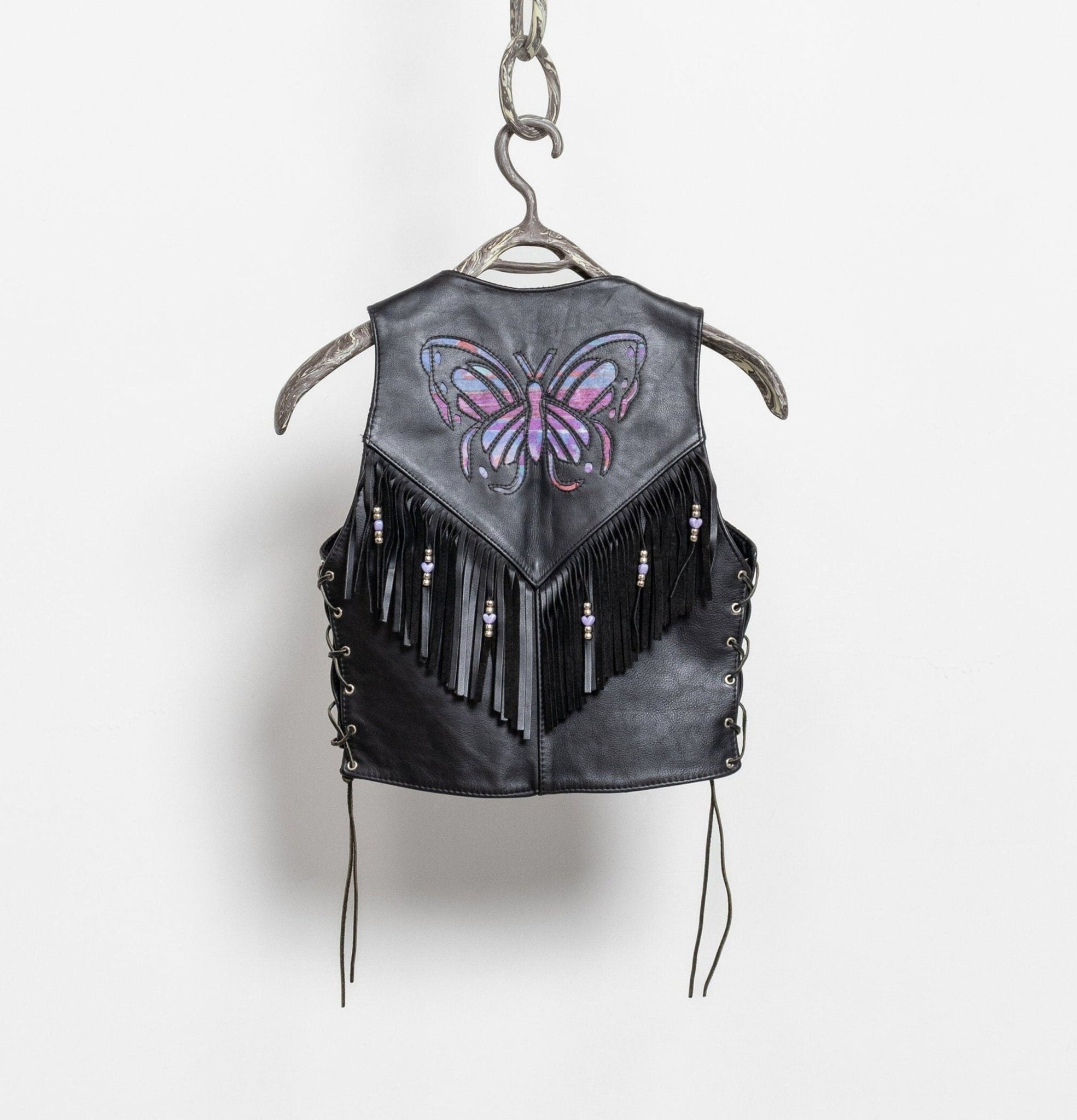 Black leather vest with fringe and butterfly design on a hanger against a white background