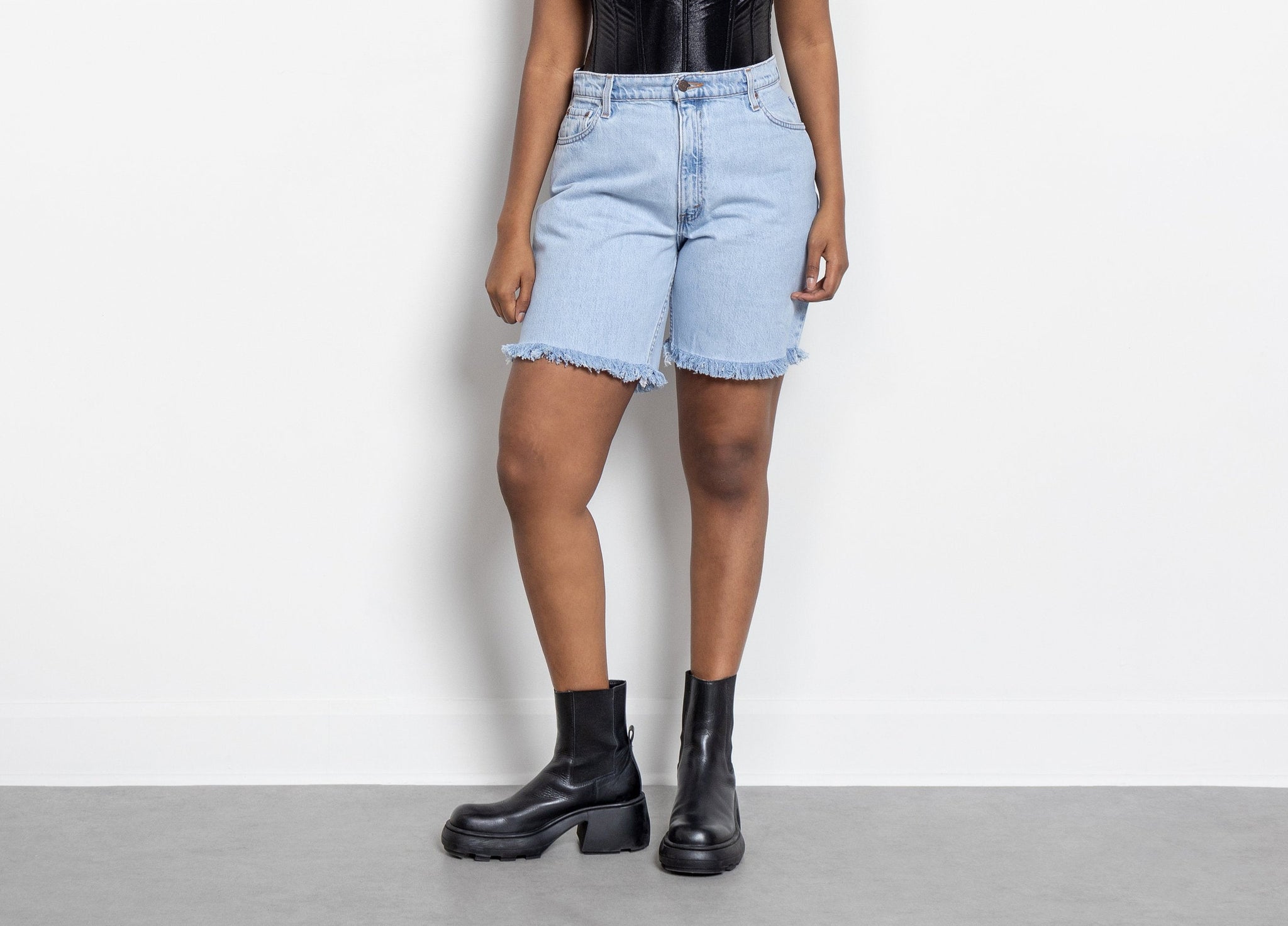 A person wearing high waist light wash frayed hem Levi's 550 longline denim shorts with black boots standing against a white background.