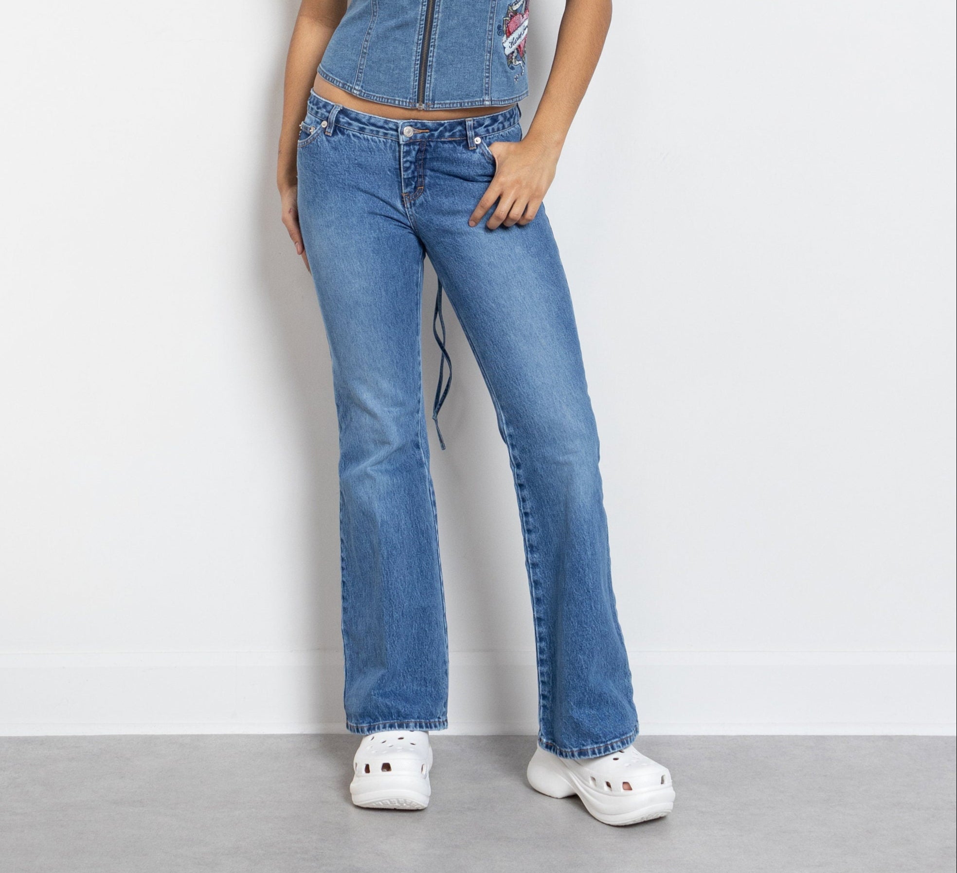 A person wearing low rise blue jeans and white shoes, standing against a plain background.