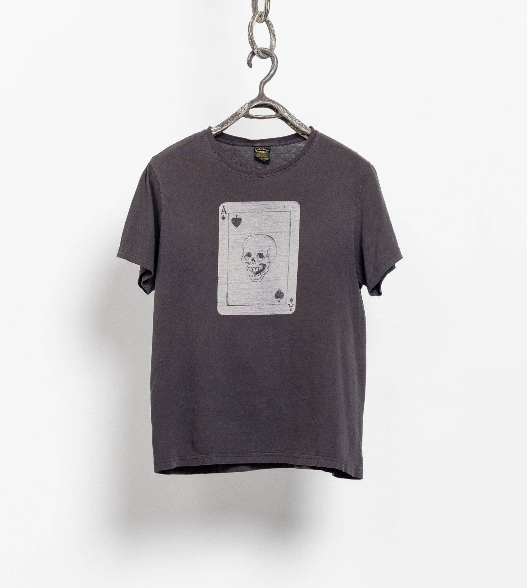 A faded black vintage t-shirt with a graphic of a skull and cards on the front, hanging from a chain against a white wall.