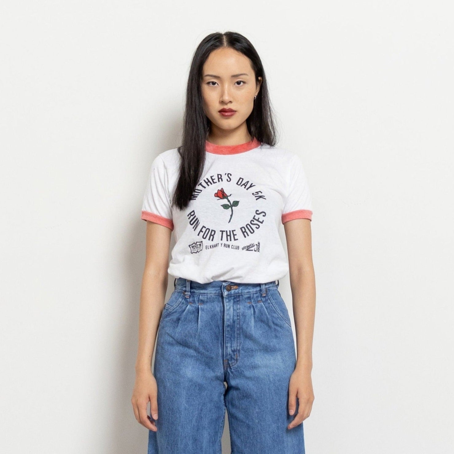 A woman wearing a white short-sleeve t-shirt with a graphic that reads 'Mother's Day 5K' and features a rose. She is also wearing blue jeans.
