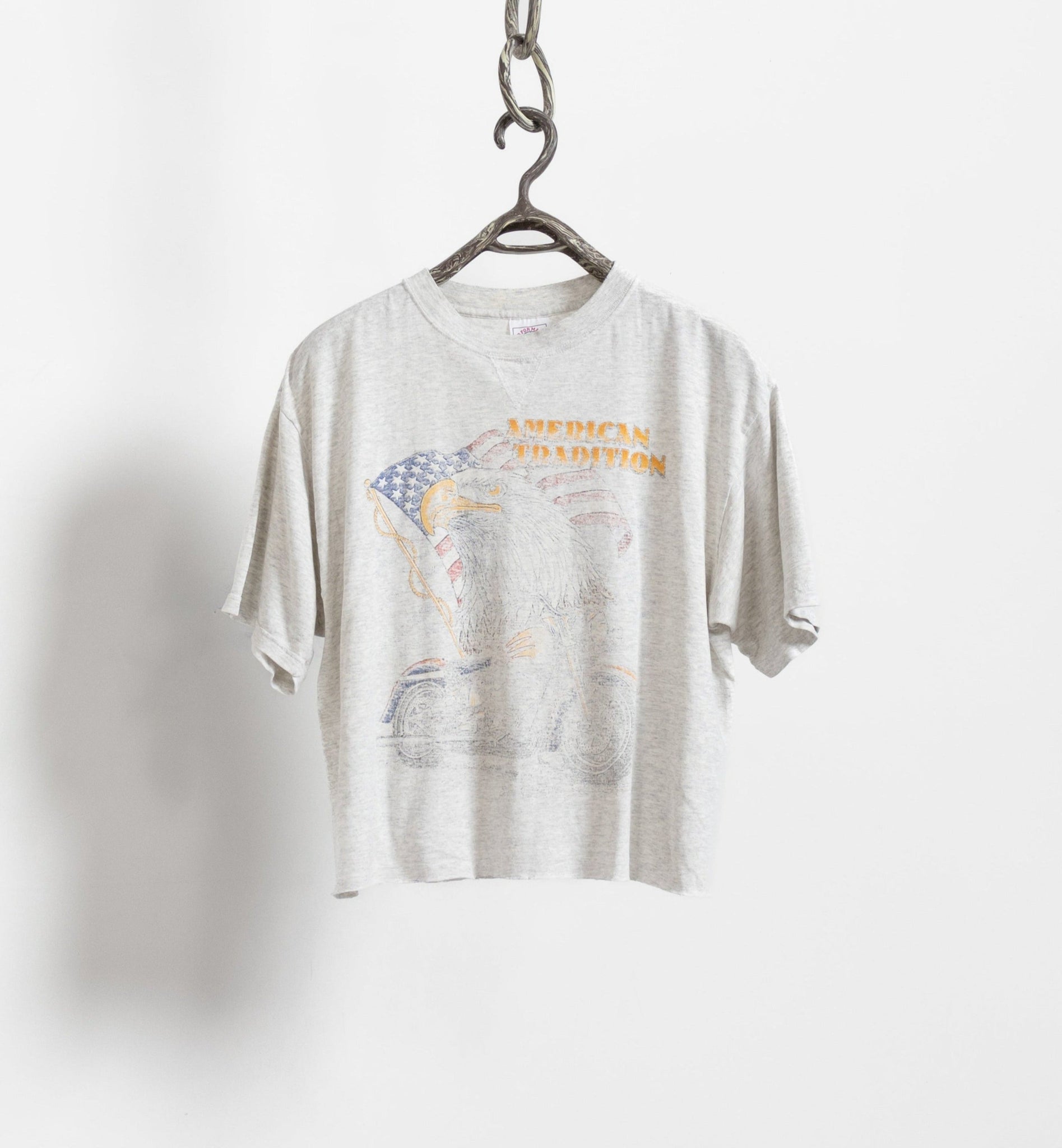 A vintage 90's cropped faded graphic t-shirt with a front eagle and motorcycle graphic, and a back graphic reading '1998 Sturgis Black Hills Rally' with an eagle on a cow skull. The shirt is beige with some wear and tear, hanging on a hanger against a white background.