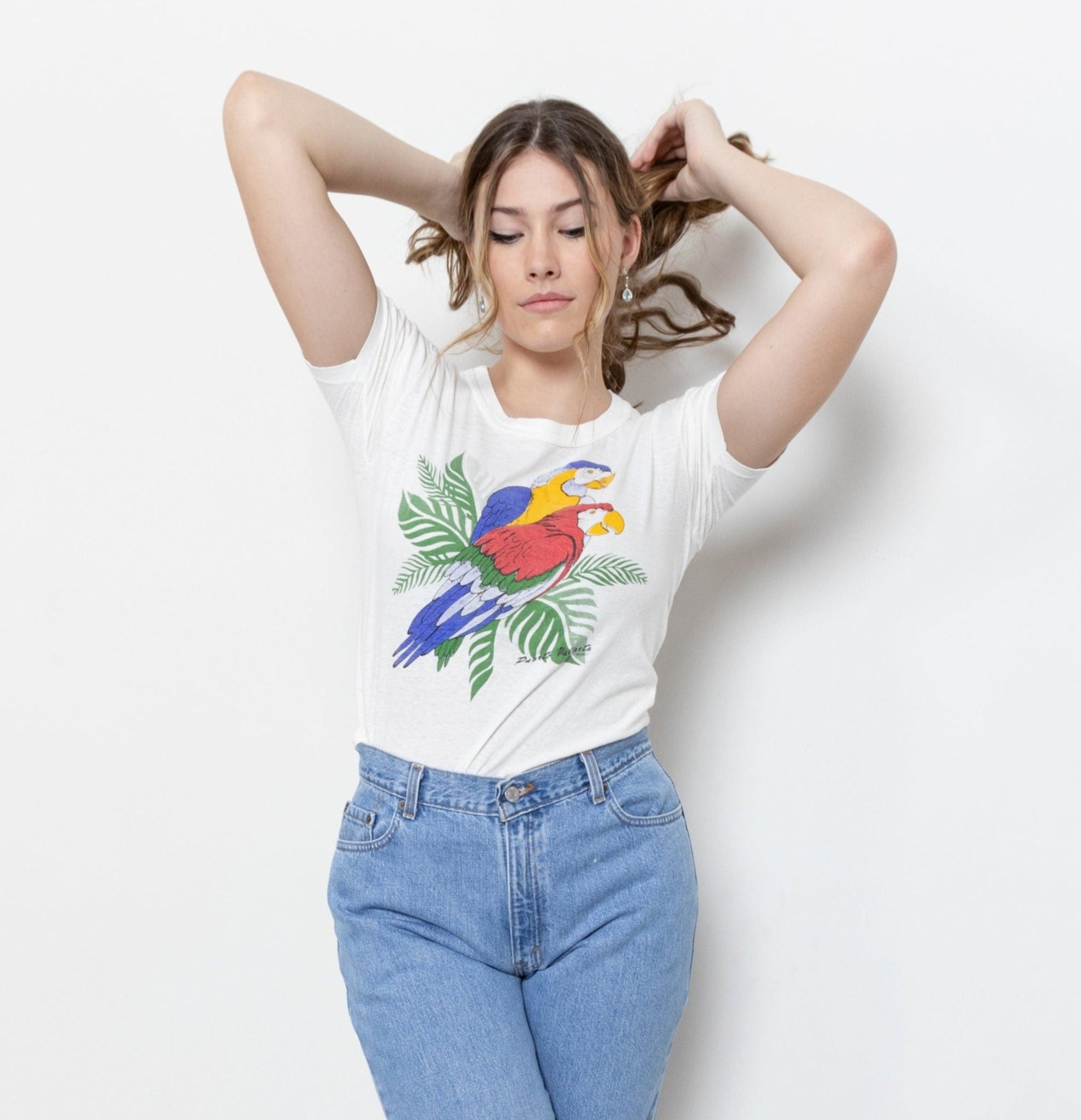 A person wearing a white short-sleeve t-shirt with a graphic of two parrots, one red and one blue, standing on a palm leaf.
