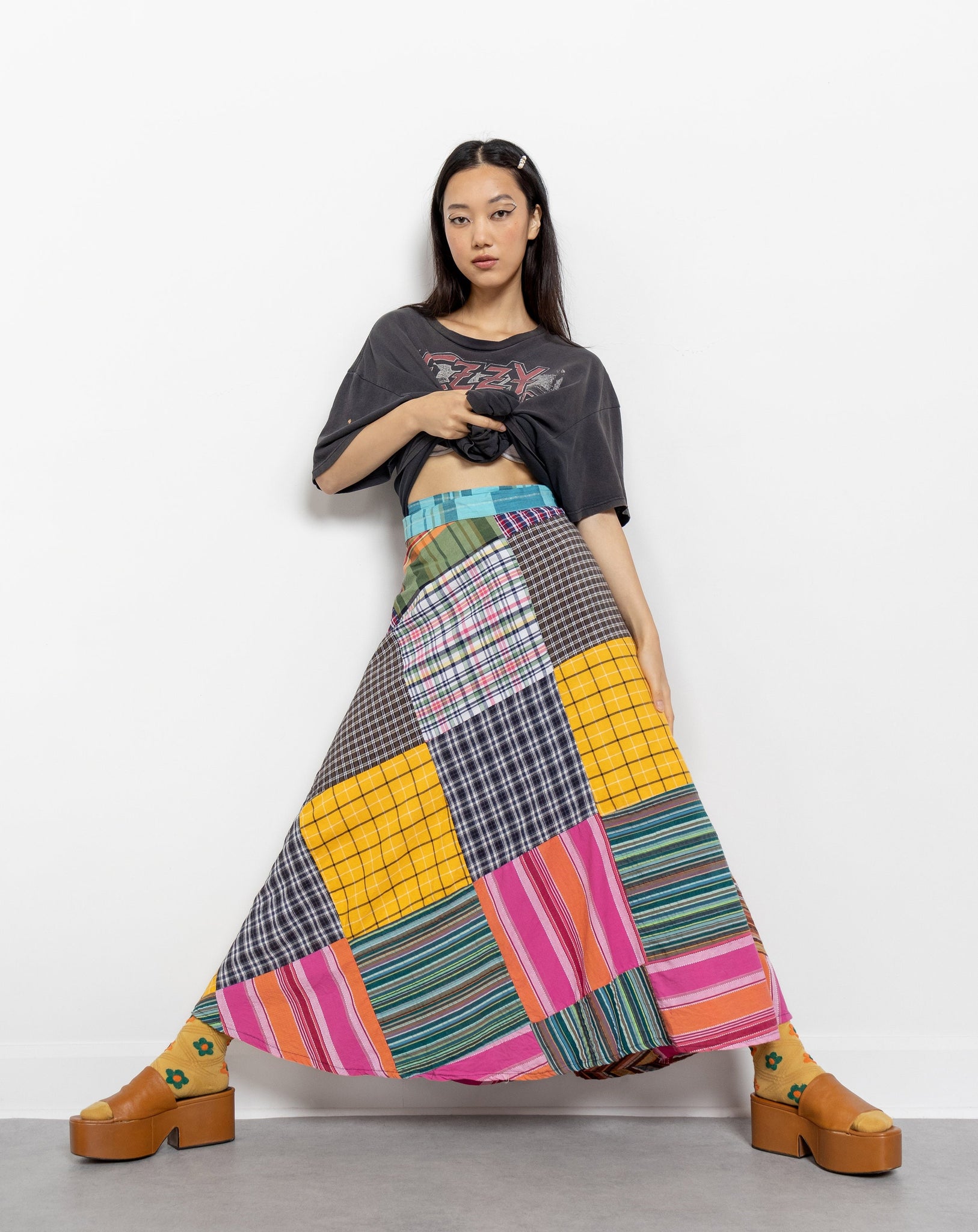 A model wearing a vintage 80's patchwork wrap maxi skirt with a bold combo of plaids and stripes in a cascade of colors. The skirt is tied up at the waist, and the model is posing against a white background.