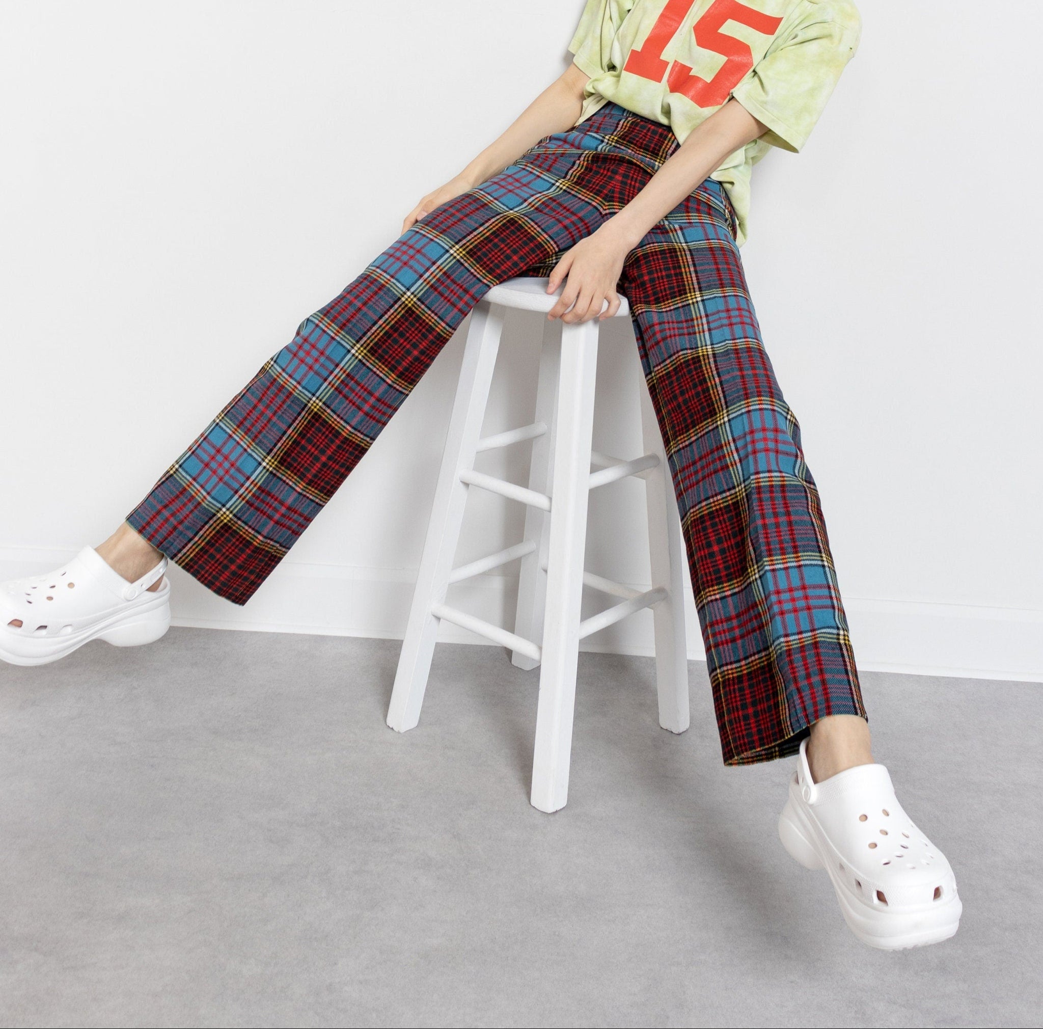 A person wearing high waisted vintage 70's plaid wool trousers in red, blue, and yellow, sitting on a white stool with a casual top and white shoes.