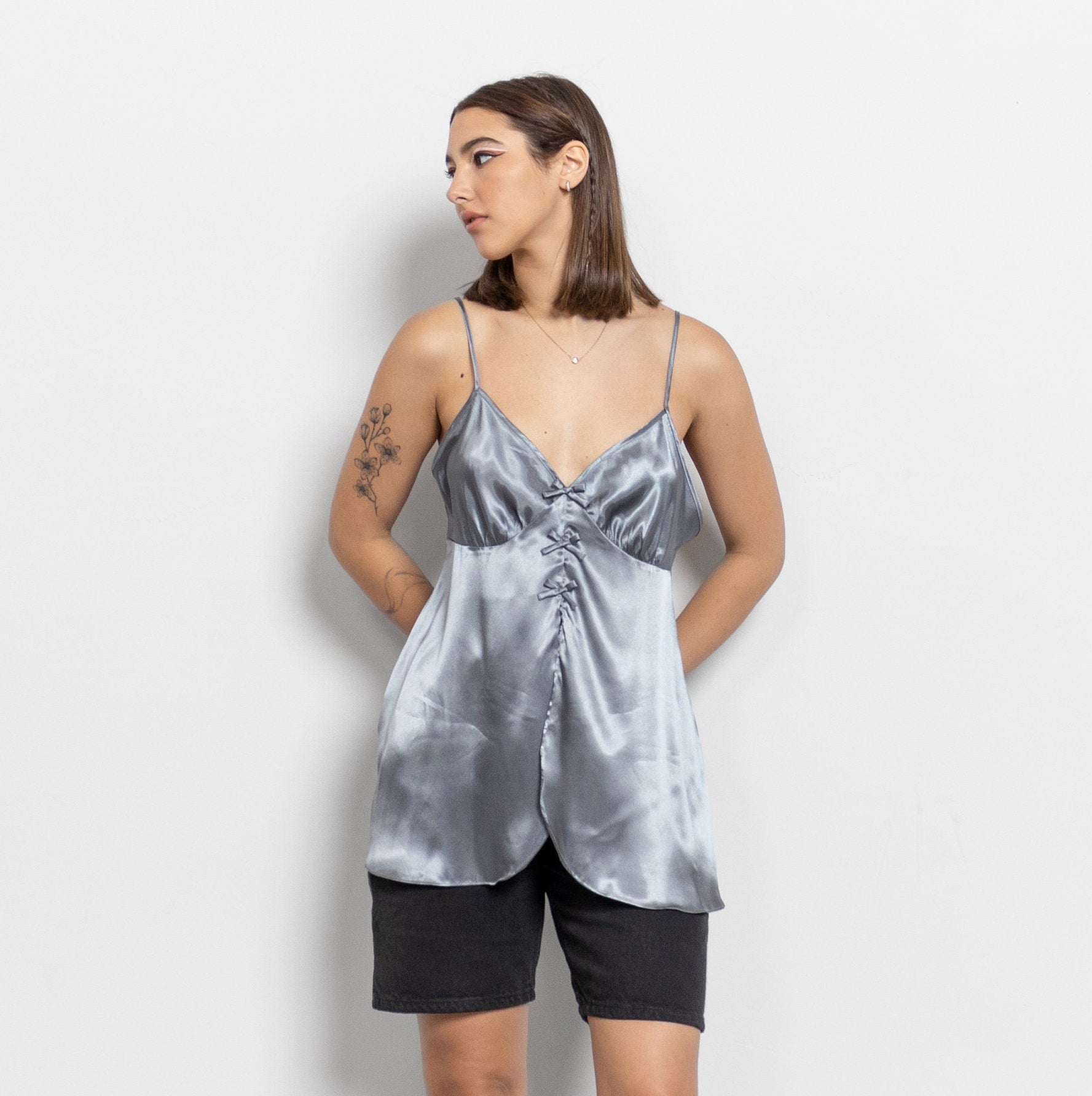 A woman wearing a silver satin camisole with spaghetti straps and a v-neckline. The camisole is flowy and has a relaxed fit with a peek-a-boo look at the front.