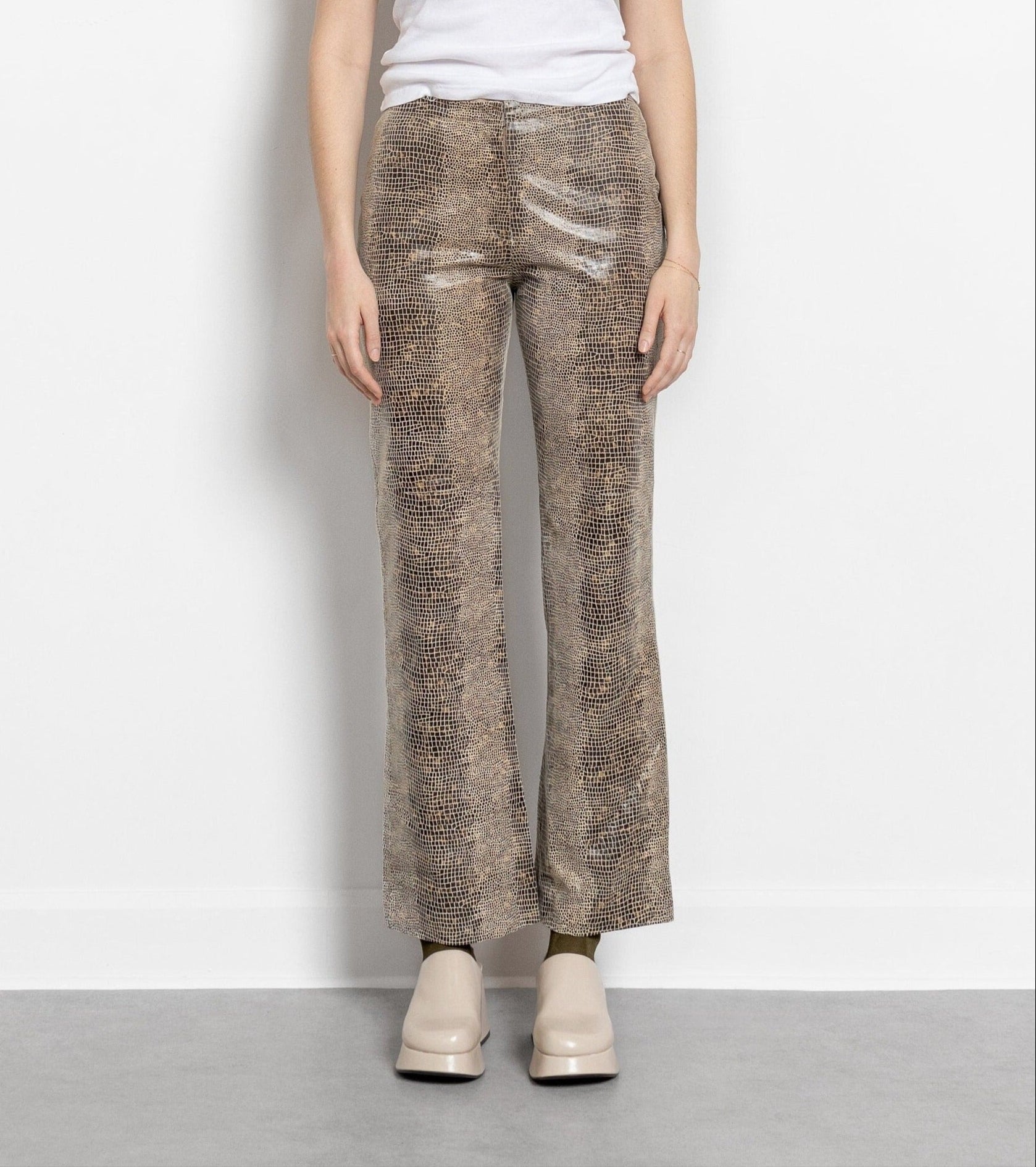 A person standing against a white background, wearing vintage 90's low rise bootcut snake print leather trousers and a white top with 'la chaussé' written on it.