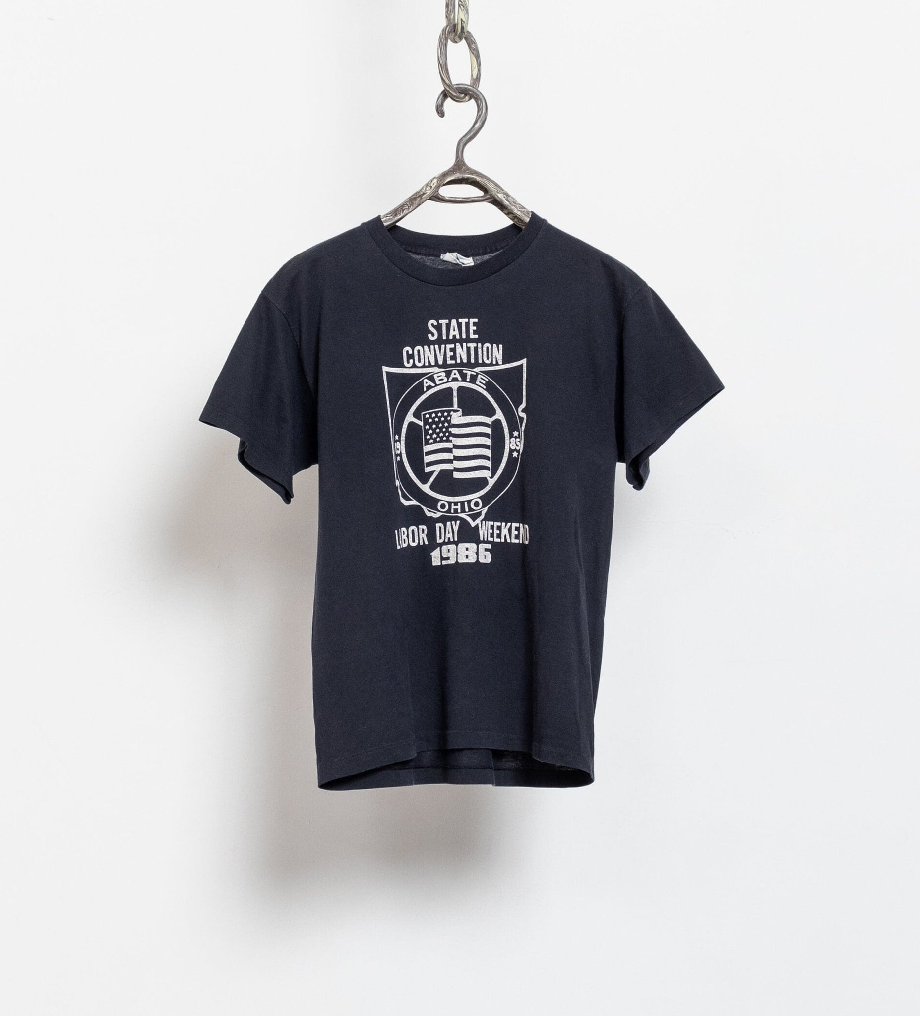 A vintage faded black crew neck t-shirt with a graphic print on the front, hanging against a white wall.