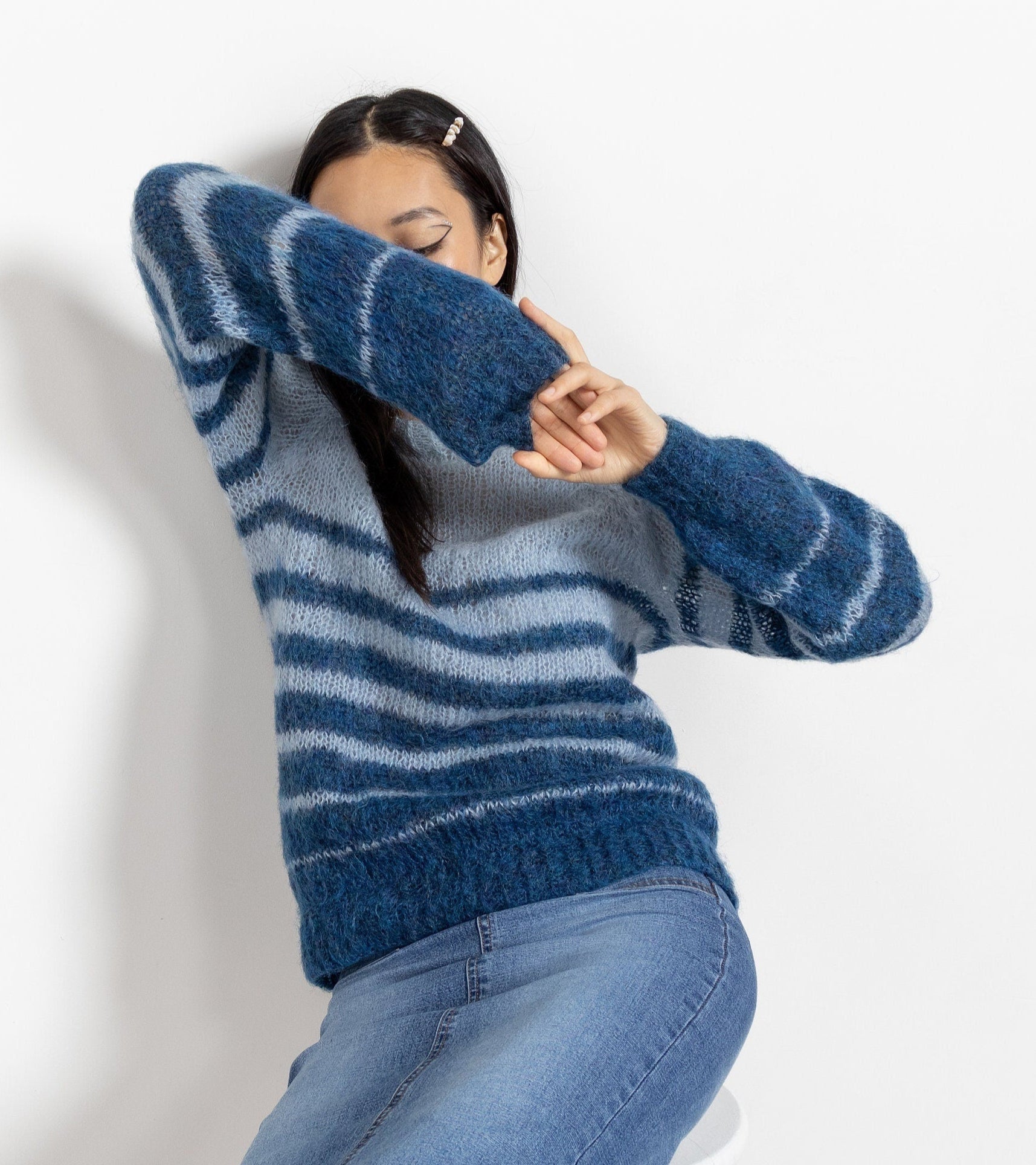 A person wearing a striped mohair-blend knit crew neck jumper in two shades of blue, seated with jeans and a green belt, against a white background.