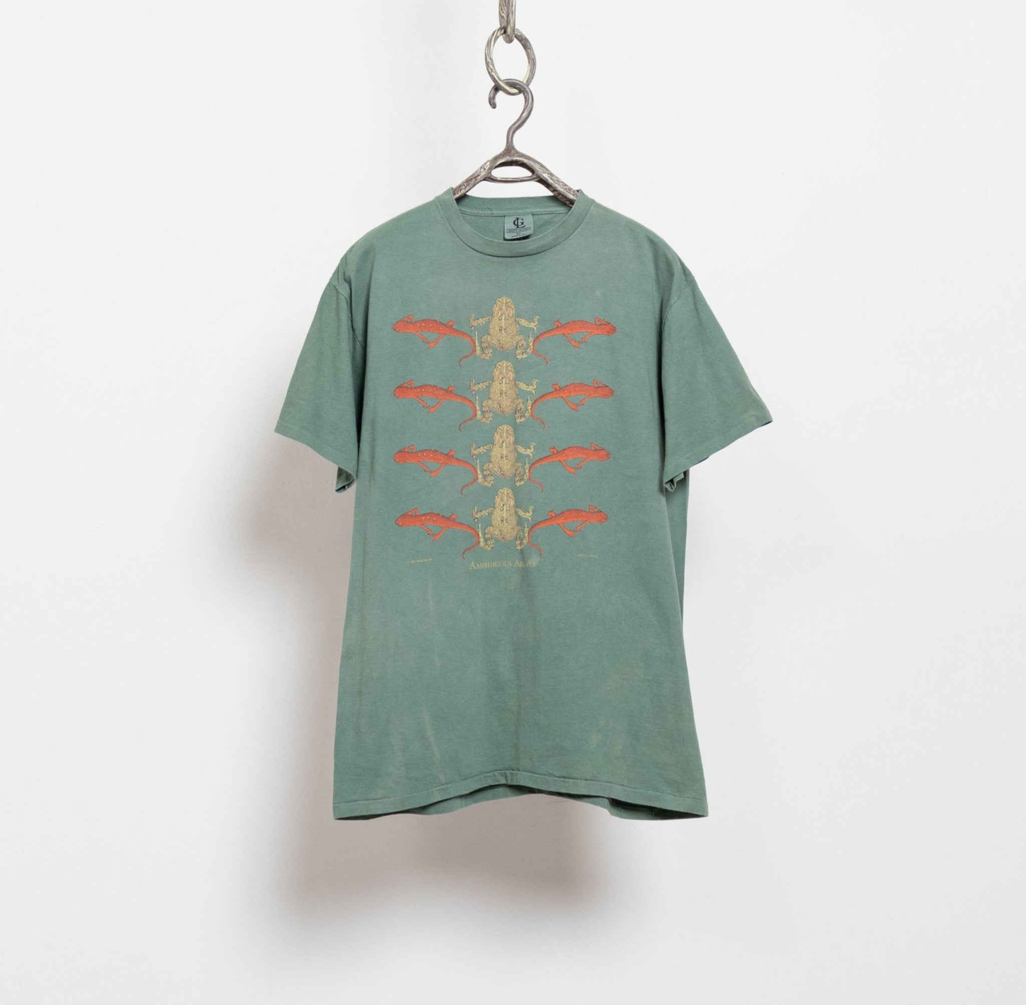 Vintage faded green t-shirt with a graphic of tadpoles and frogs in shades of orange and yellow, hanging on a hanger against a white background.