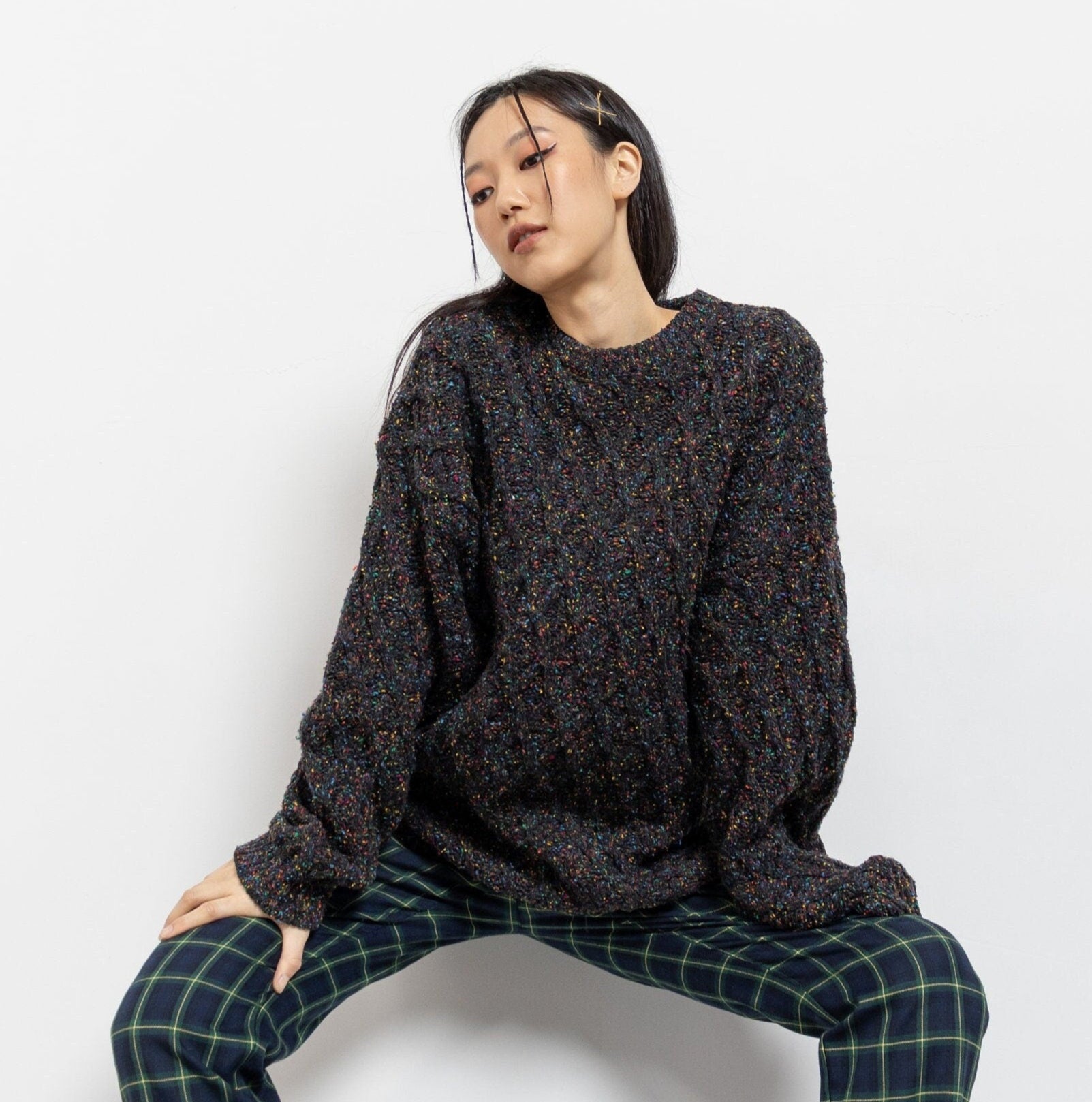 A person wearing a dark gray or black cable knit wool-blend pullover sweater with rainbow flecks, paired with checkered pants.
