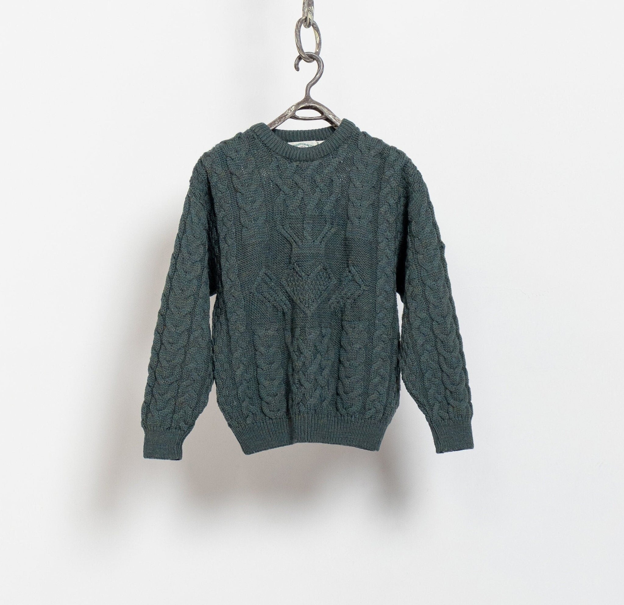 A green cable knit Aran Crafts pullover sweater hanging against a white background.