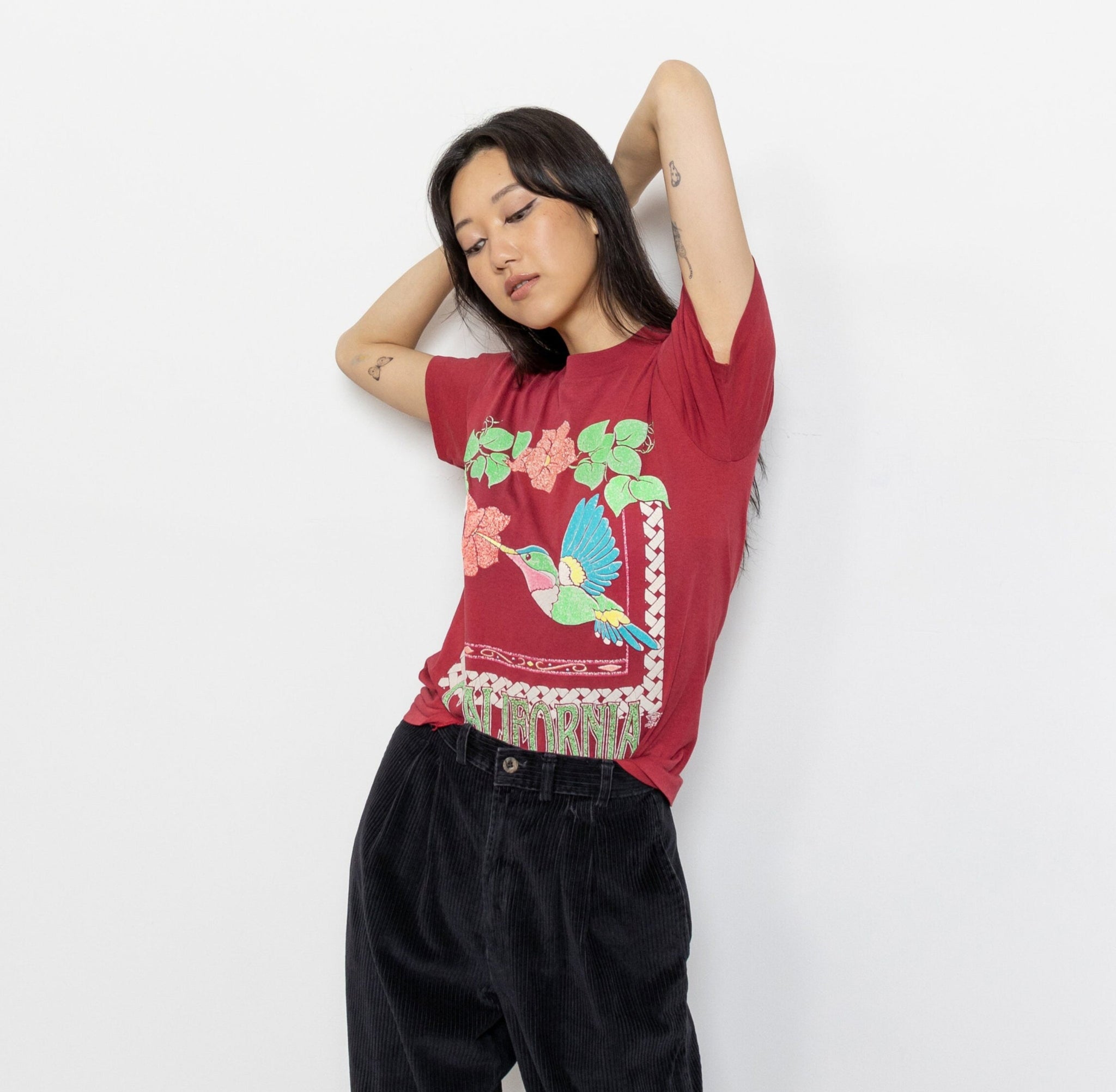 A person modeling a vintage 80's maroon California t-shirt with a graphic of hummingbirds and text, paired with black pants.