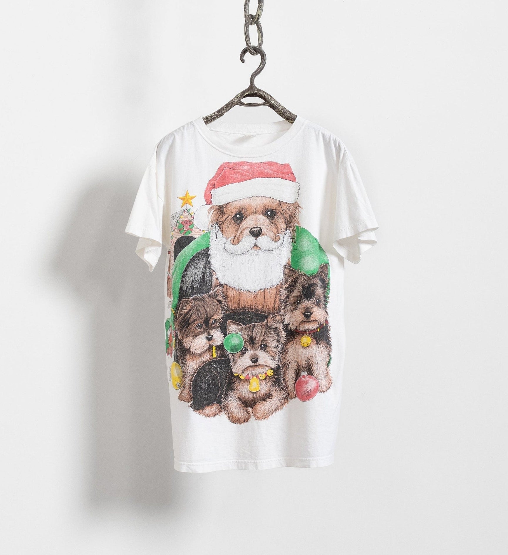 A white crew neck t-shirt with a graphic print of a dog dressed as Santa Claus and surrounded by small dogs, decorative elements like stars and Christmas trees, and text reading 'Holiday Doggies'.