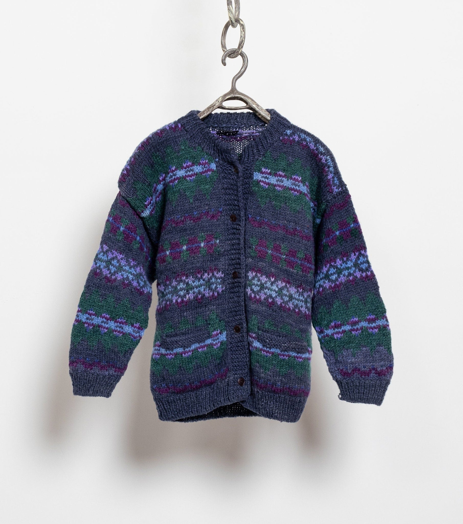 A vintage 90's chunky hand knit oversize cardigan sweater with a bold fair isle pattern in purple and blue / black colors, hanging on a chain against a white wall.