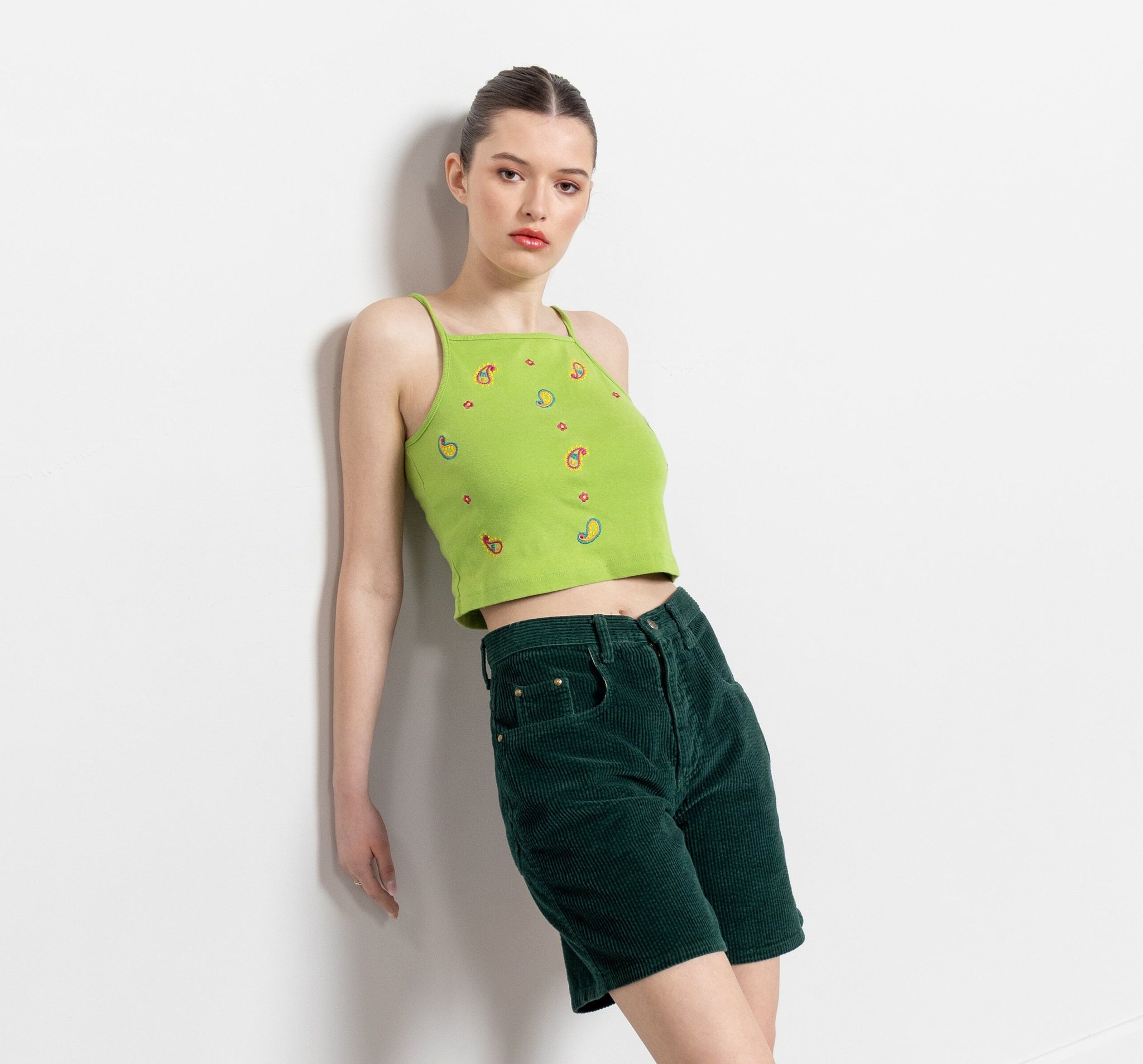 A woman wearing vintage green corduroy high-waisted shorts and a matching green top with a floral pattern, posing against a white background.