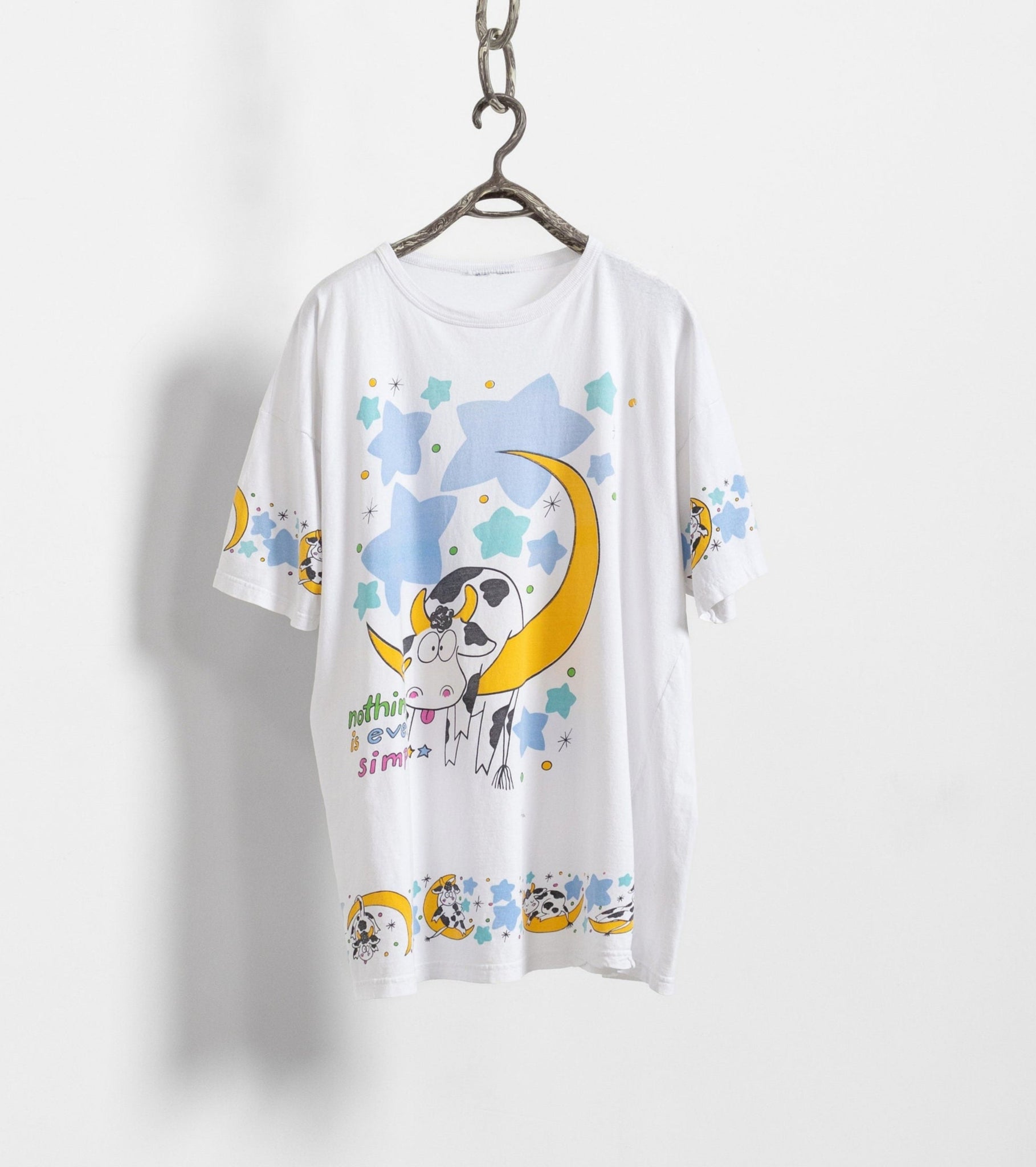 An oversized white t-shirt with a graphic print of a cow, moon, and stars in pastel colors, displayed on a hanger against a white background.