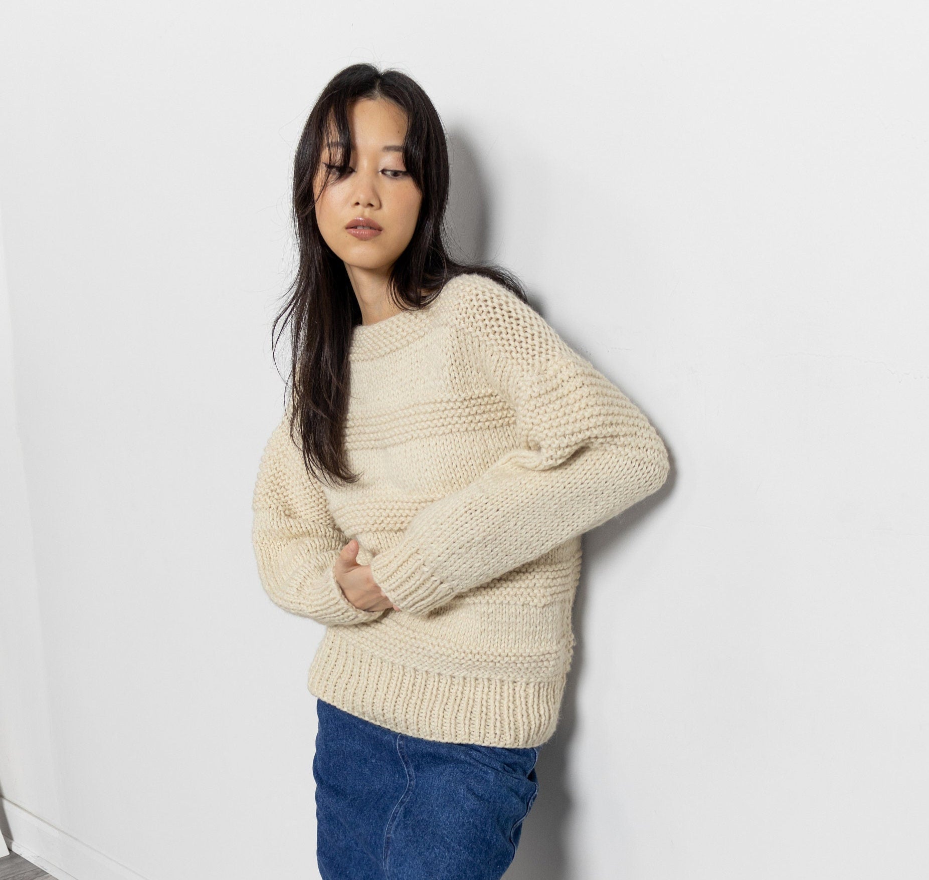 Woman wearing a beige sweater, blue skirt, and white boots against a white wall.