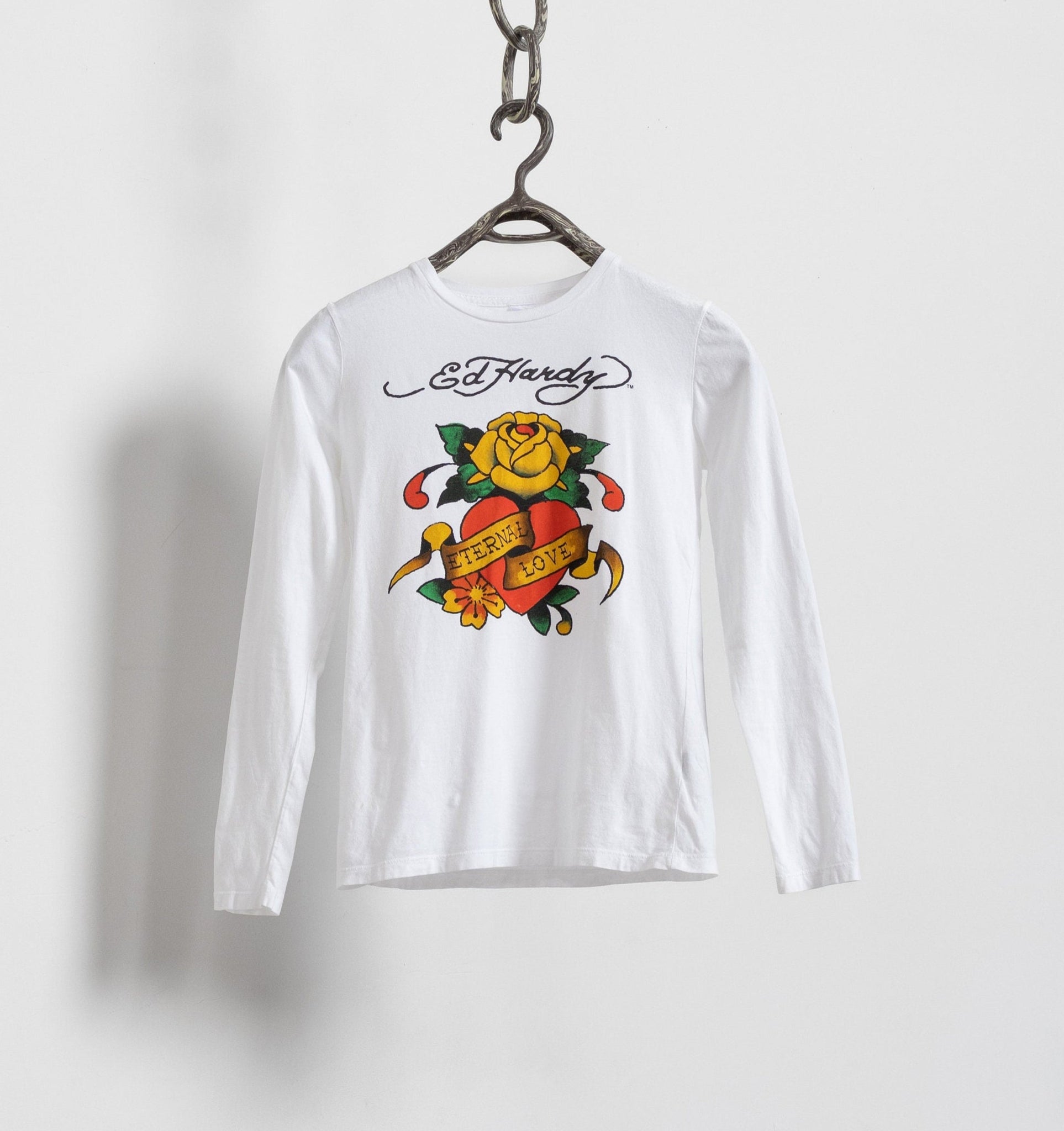 A white long sleeve Ed Hardy t-shirt with a black and white line work of a classic rose and heart motif and the words ETERNAL LOVE scrawled across the front, hanging on a chain.