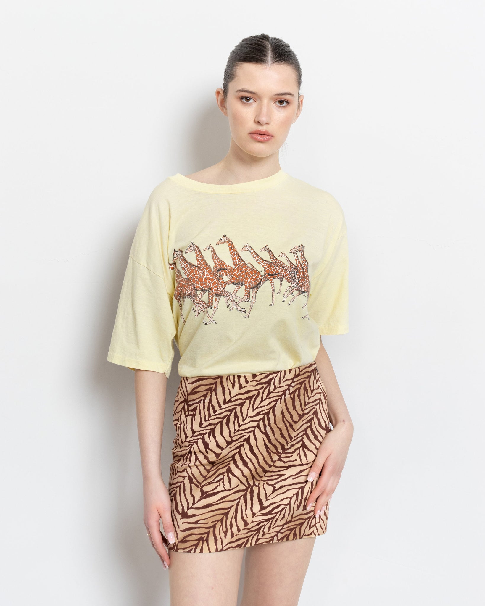 A person wearing a vintage pale yellow cotton t-shirt with a Chicago field museum graphic, featuring giraffes, and a brown skirt with a animal print.