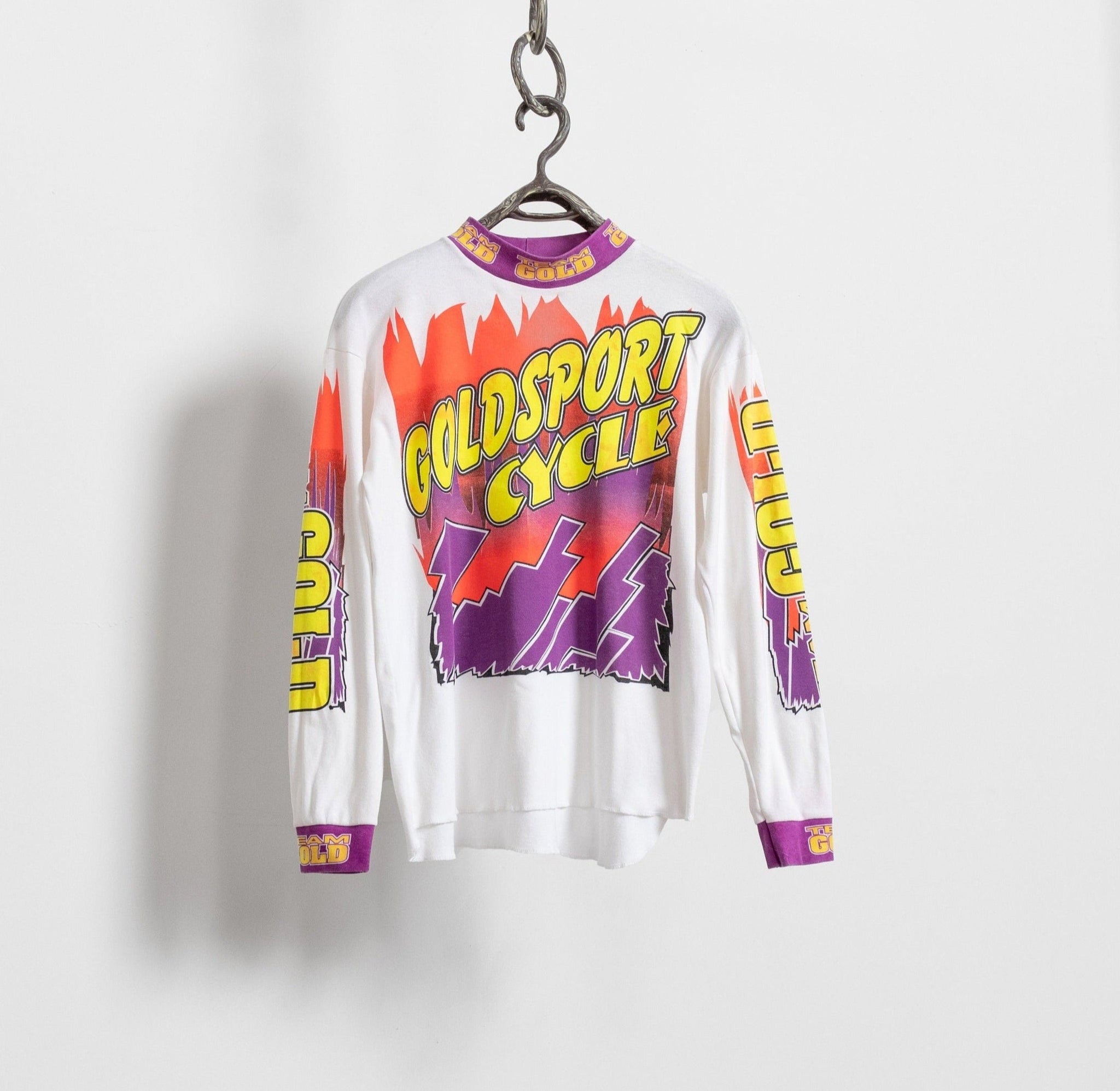 A long sleeve white cotton Goldsport Cycle mockneck t-shirt with bold pink and purple graphics and yellow letters, displayed on a hanger against a white background.
