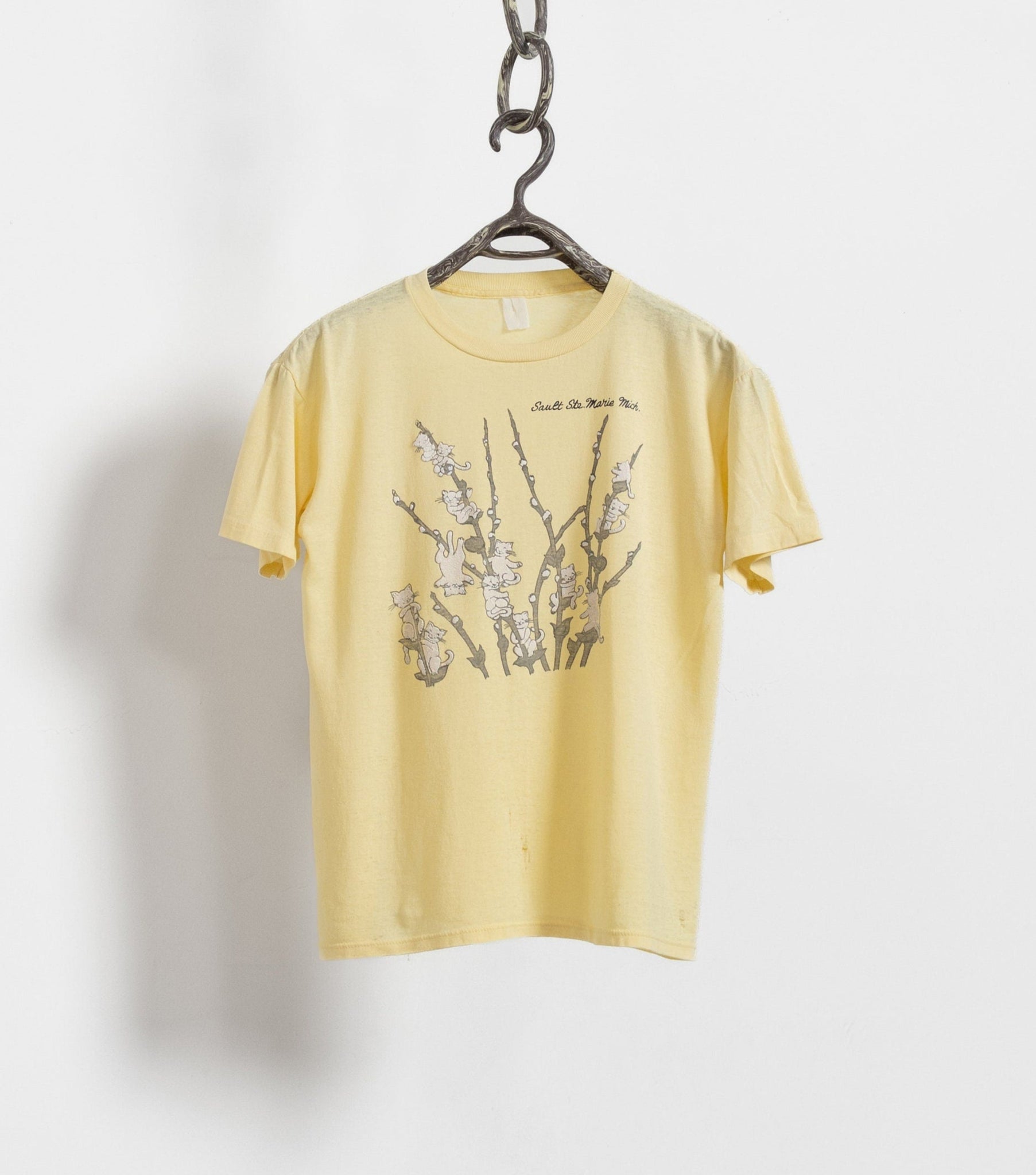 A yellow short-sleeve t-shirt with a graphic print of cats on branches, hanging on a hanger against a white background.