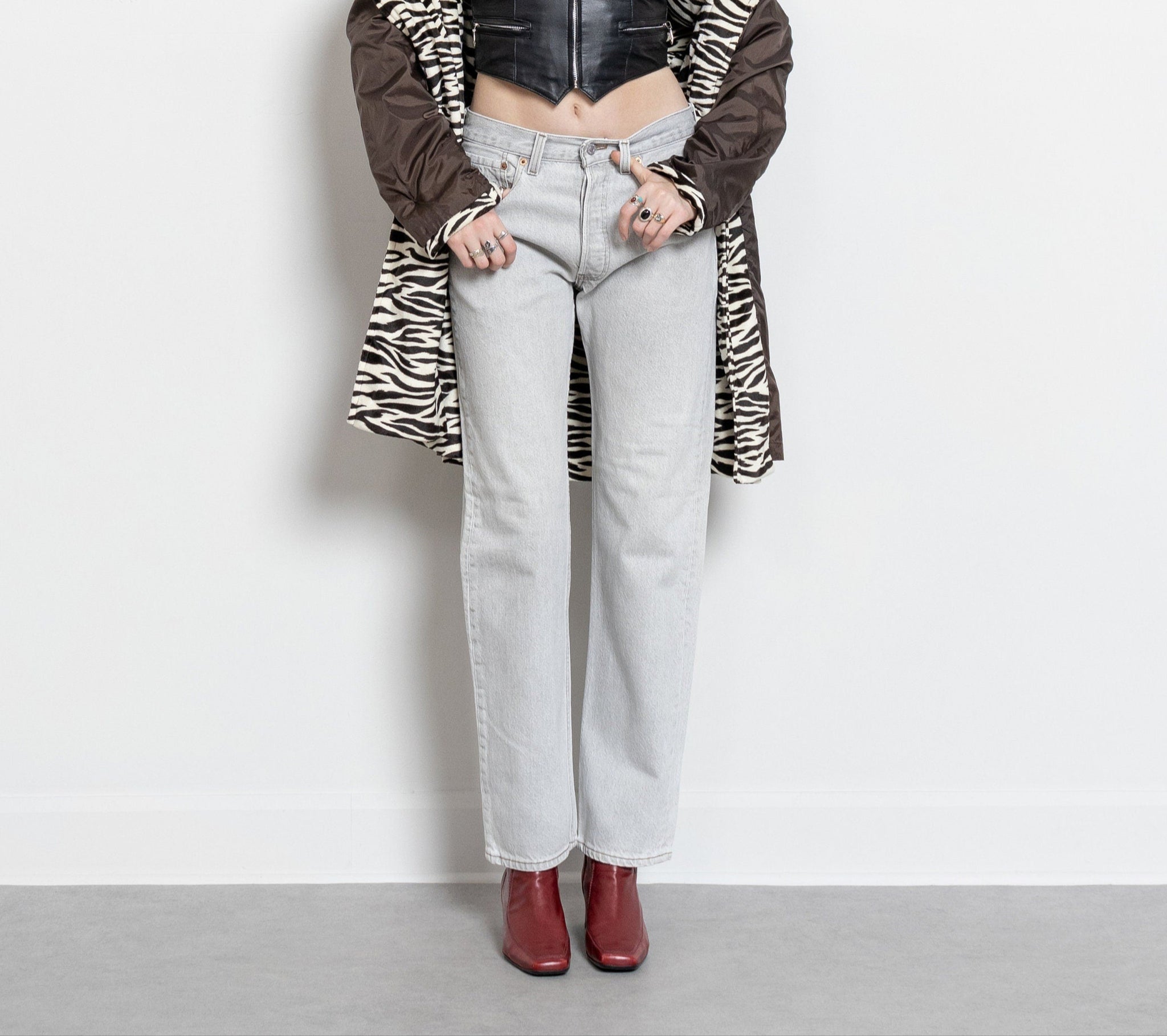 A person standing against a white background, wearing vintage Levi's 501 jeans with a button fly, light grey in color, with brownish hues. They are also wearing a zebra-patterned coat and red boots.