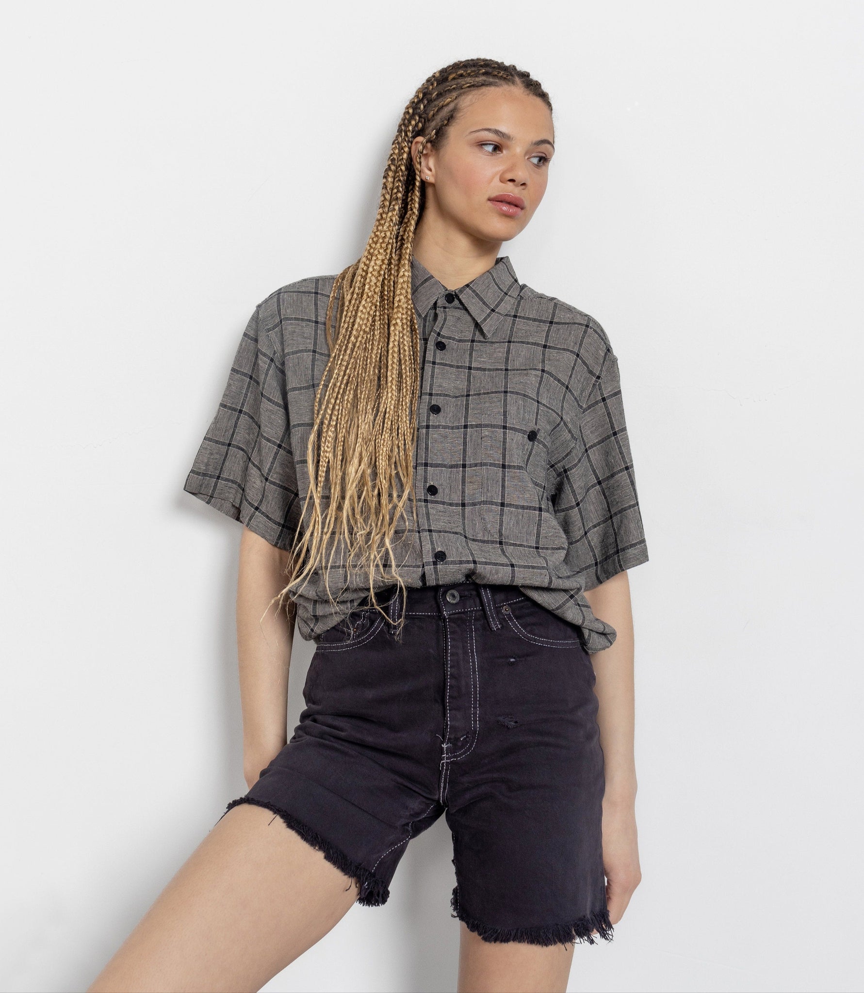 A female model wearing a plaid short-sleeved button-up shirt and high-waisted black cut-off denim shorts.