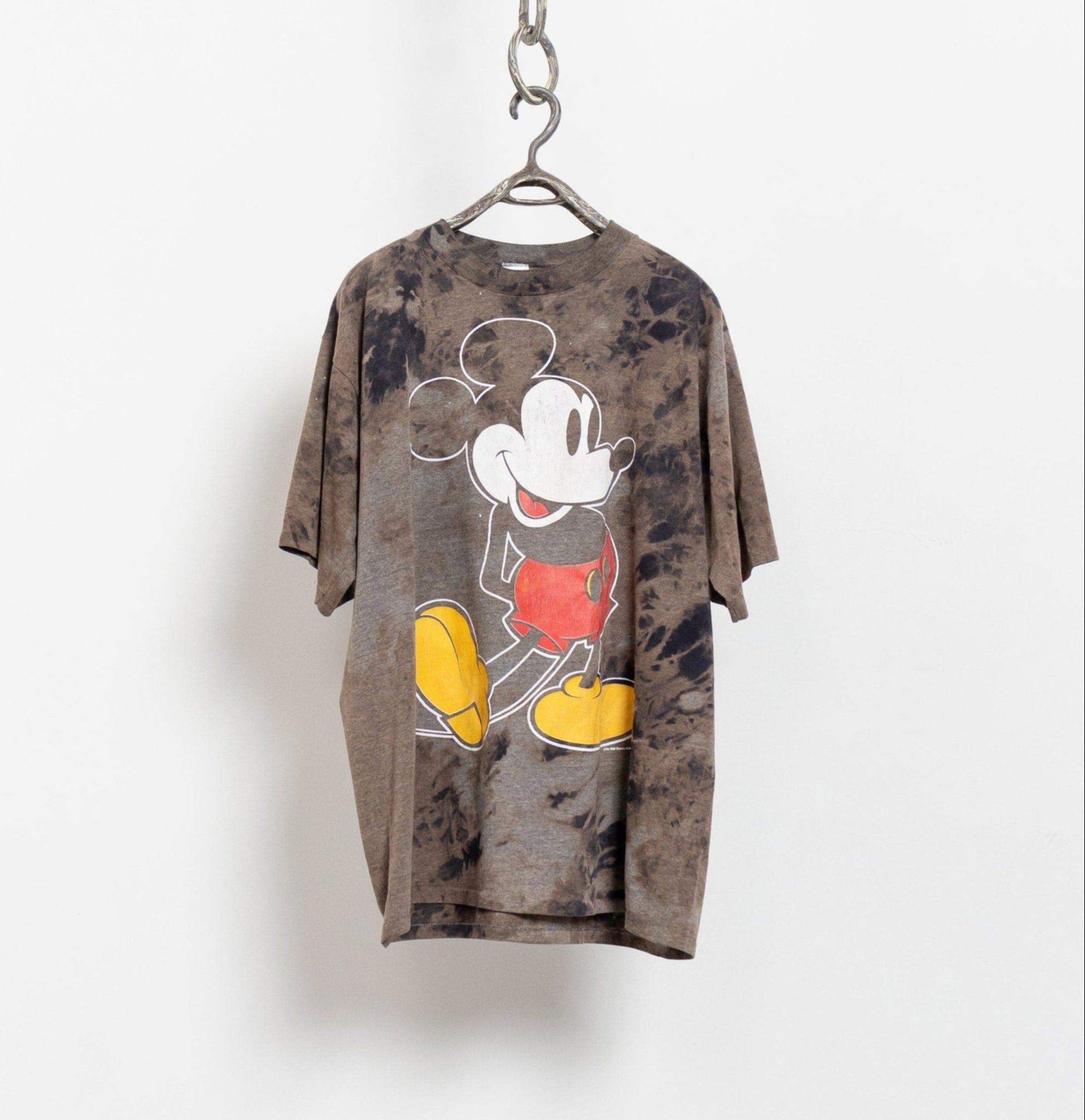 An oversized, tie-dye t-shirt featuring a vintage Mickey Mouse graphic in red and yellow on a dark background, hanging against a white backdrop.