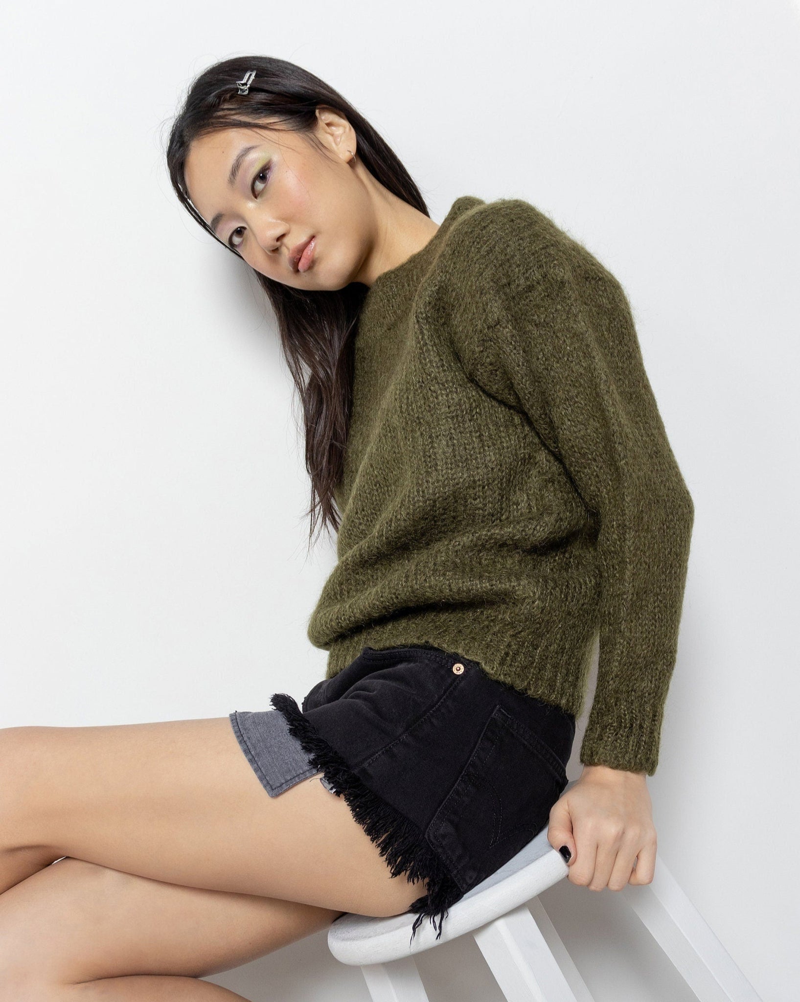 A woman sitting on a stool wearing an olive green, mock-neck pullover sweater with a slightly cropped length.