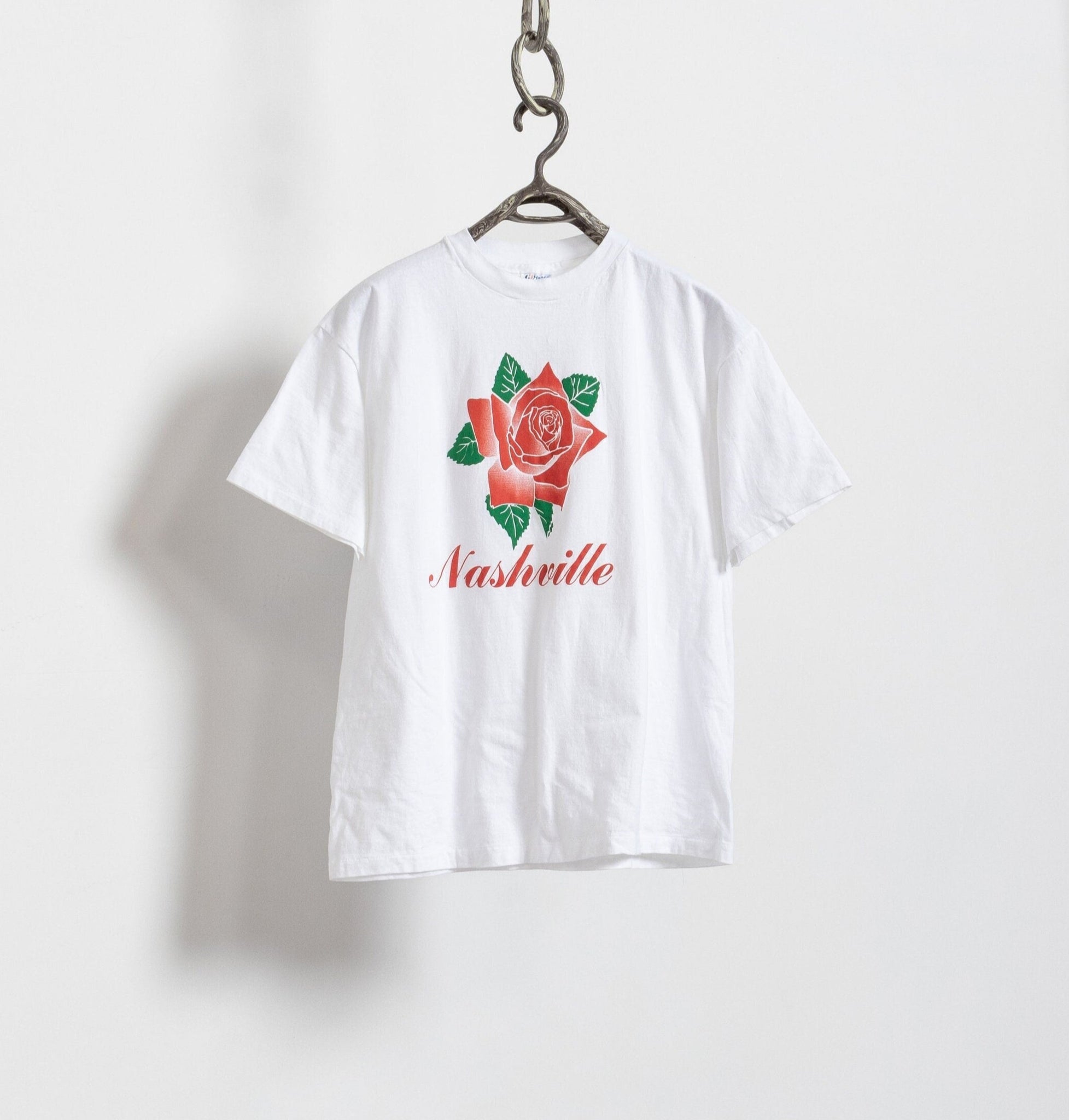A white vintage 90's graphic tee with a red rose and the word 'NASHVILLE' printed on the front, hanging against a plain background.