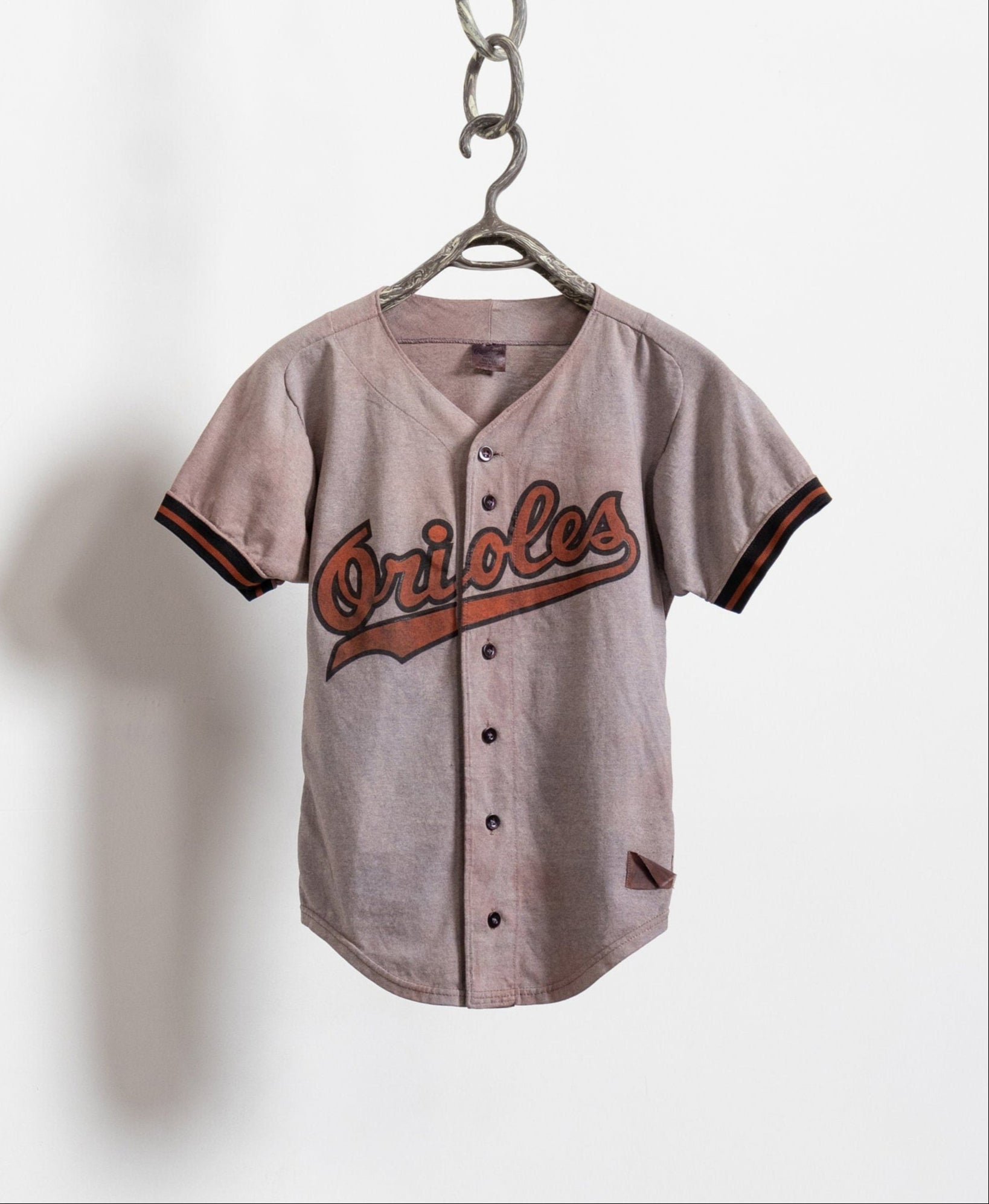 A vintage 80's Orioles button up poly cotton baseball tee in tie-dyed brown, with the team's number 1 on the back, hanging on a chain against a white wall.