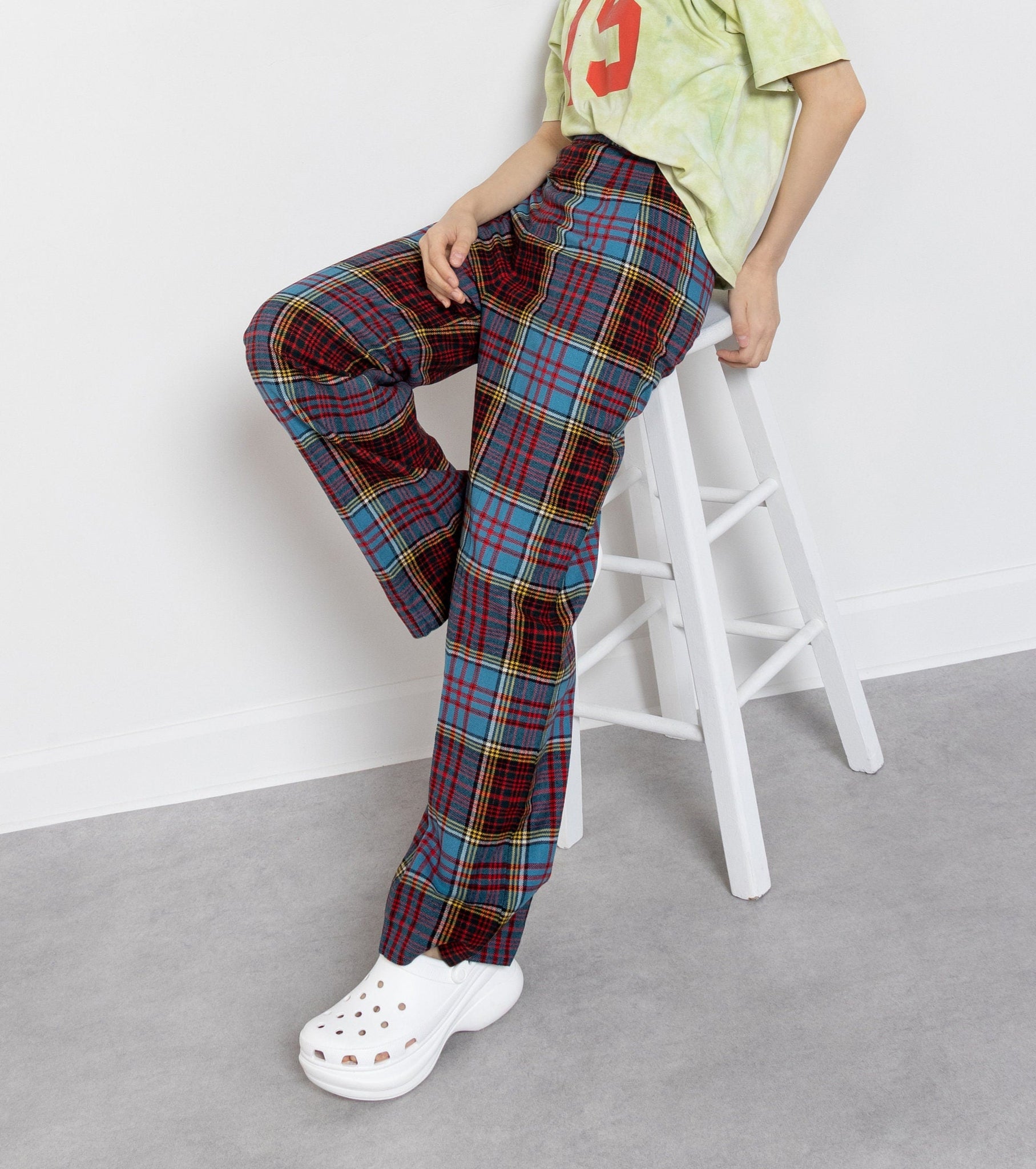 Woman wearing a light green t-shirt with a red number and plaid pants sitting on a white stool against a white wall.