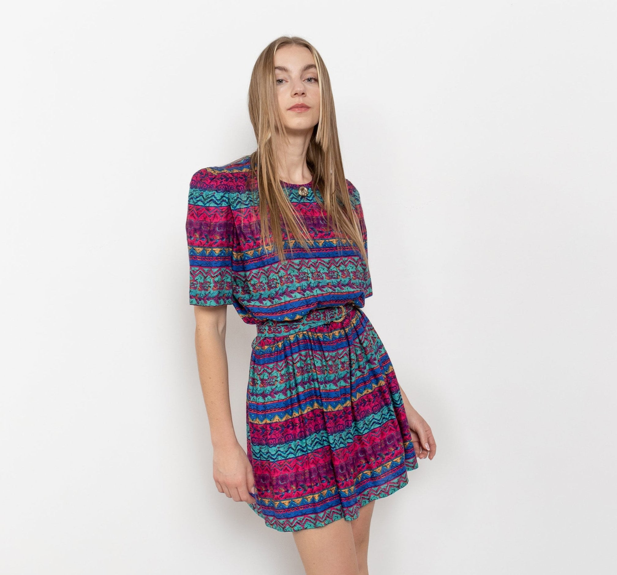 A model wearing a vintage 90's colorful abstract print short sleeve crew neck mini dress with an elastic waist, shoulder pads, and side pockets.