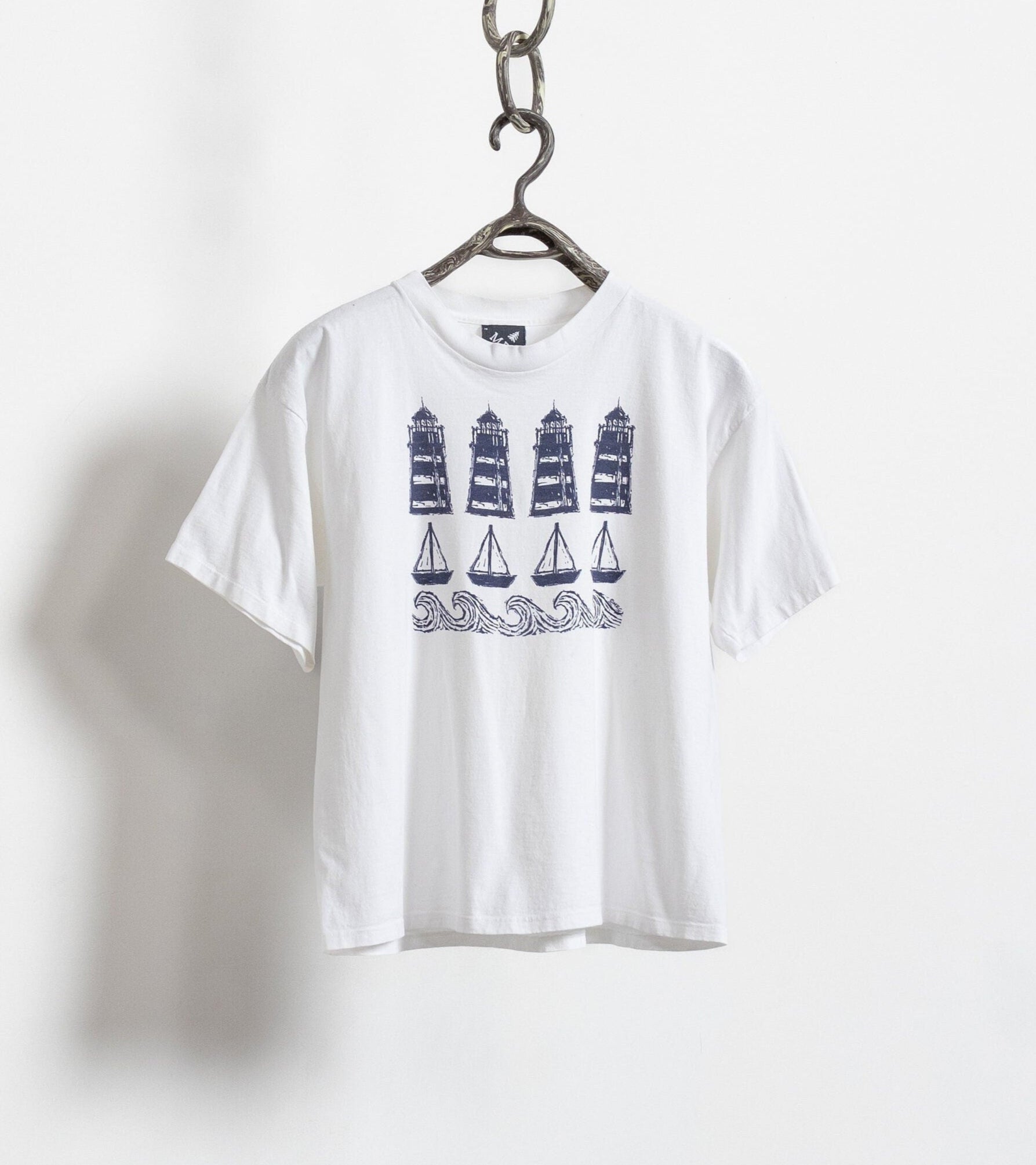 A white, oversized t-shirt with a vintage lighthouse print graphic, hanging on a chain against a white wall.