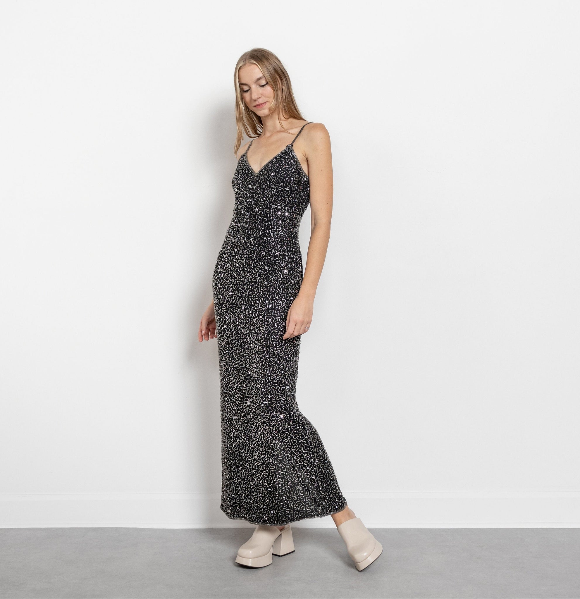 A woman modeling a vintage black silk sequin maxi dress with spaghetti straps and a V-neckline. The dress is embellished with sequins and beads, has a relaxed silhouette, and zips up the back.