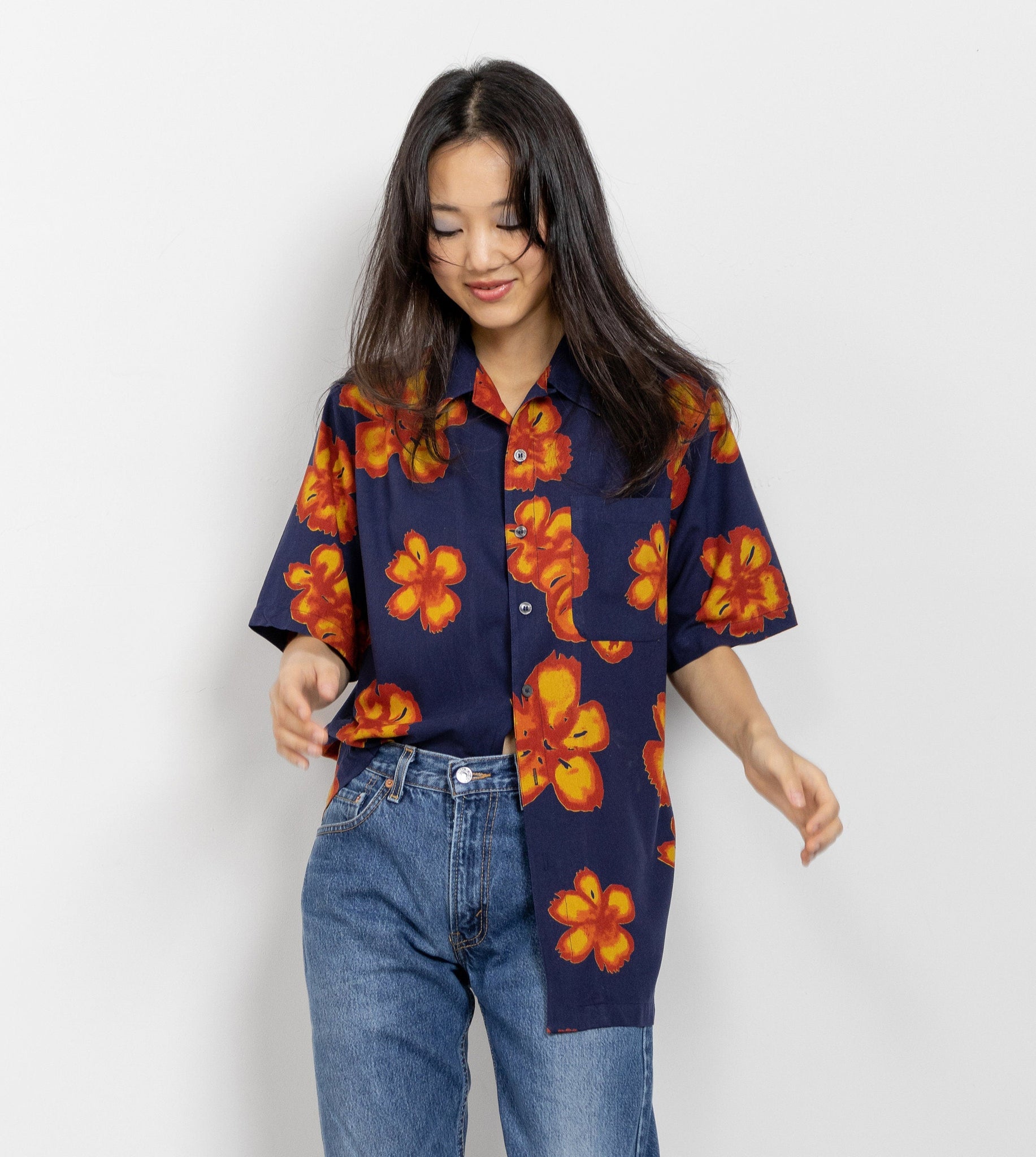 A person wearing a short-sleeved navy blue silk Hawaiian shirt with a floral pattern of glowing orange tropical hibiscus flowers, paired with blue jeans.