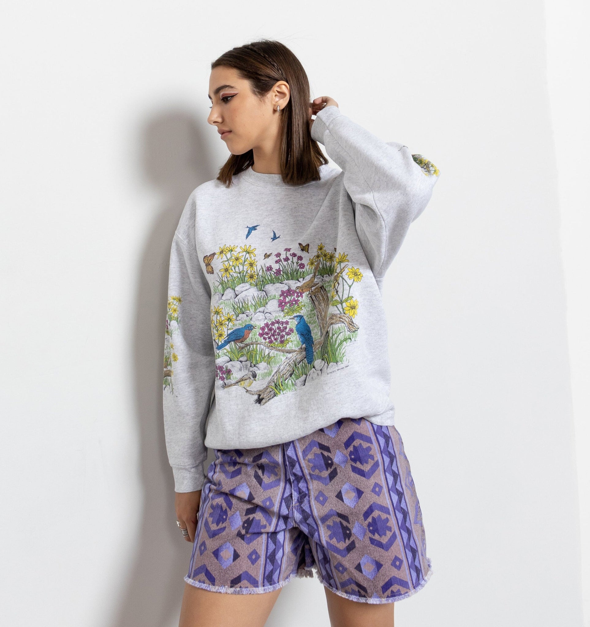 Woman wearing a gray sweatshirt with a colorful design and purple patterned shorts on a white background