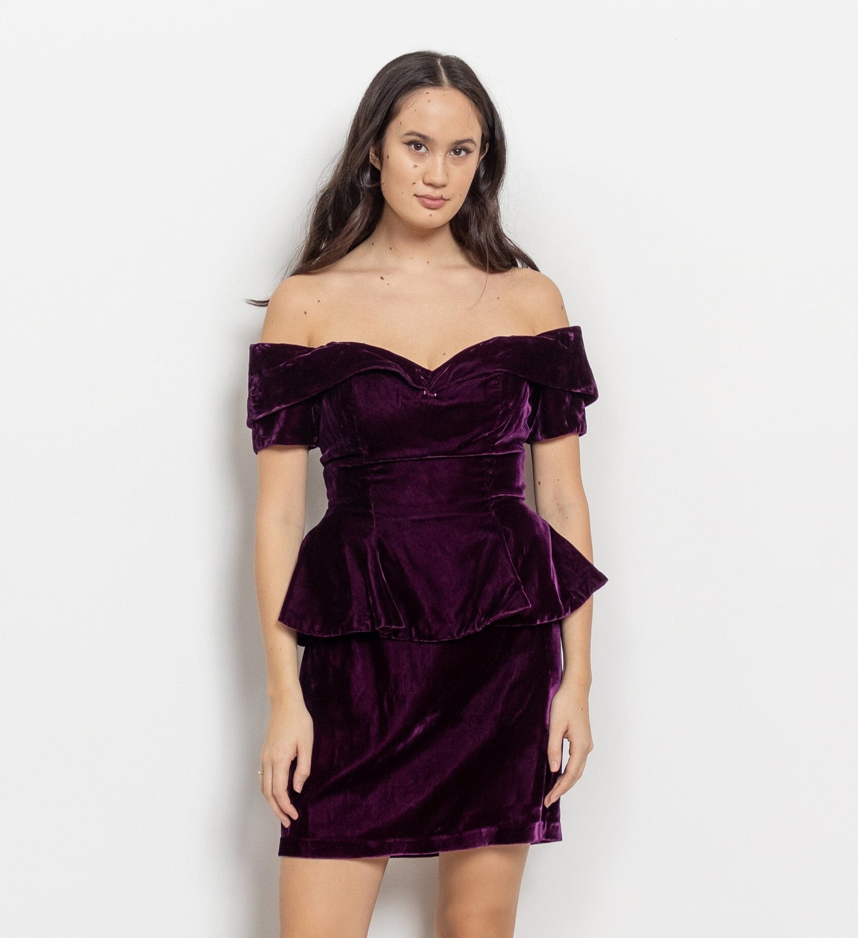 A model wearing a vintage 90's purple velvet off the shoulder mini dress with short sleeves and a peplum waist.