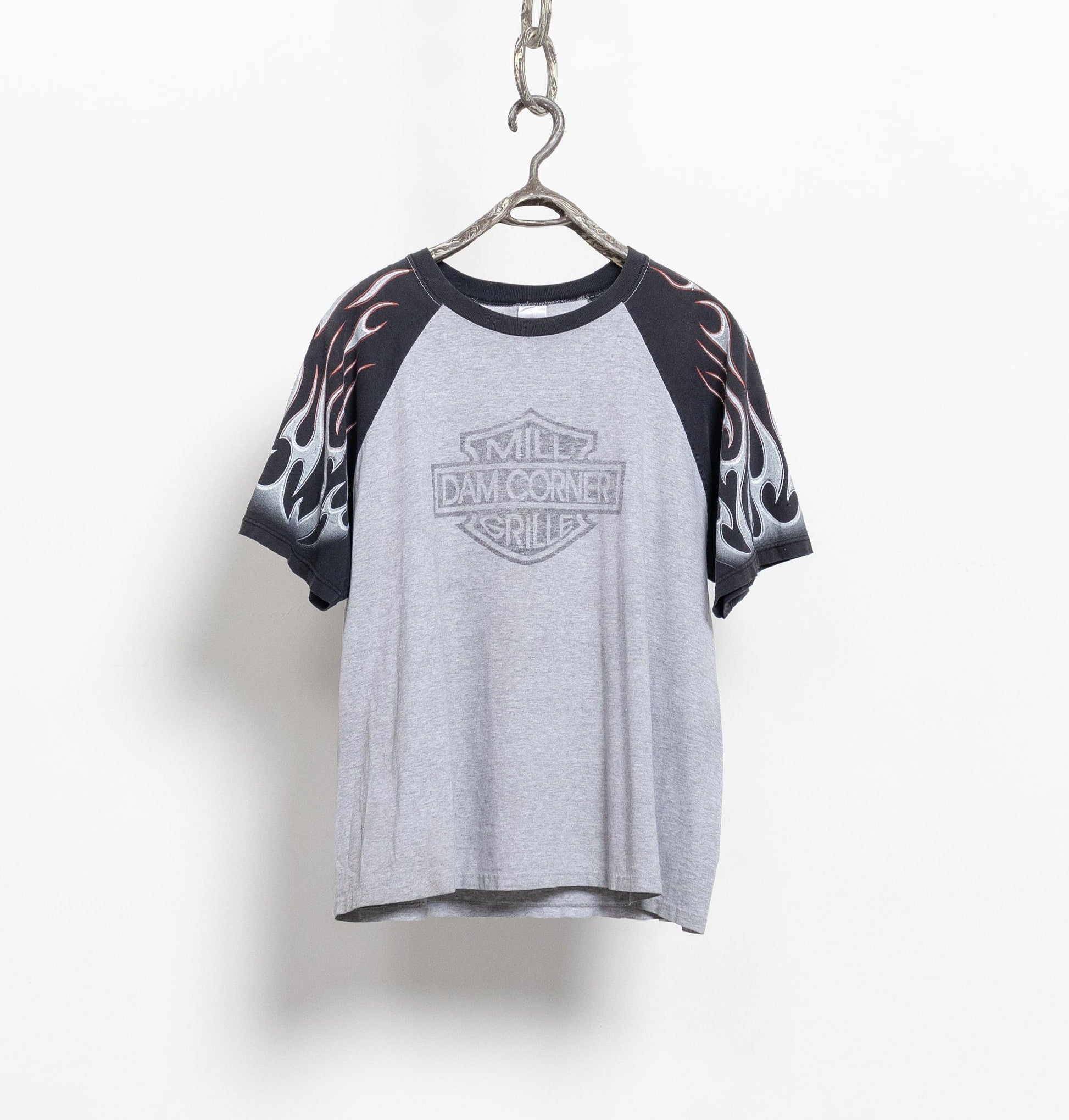 A vintage 90's style Mill Dam Corner Grill restaurant graphic tee in heather grey with black sleeves and trim, hanging against a white wall.