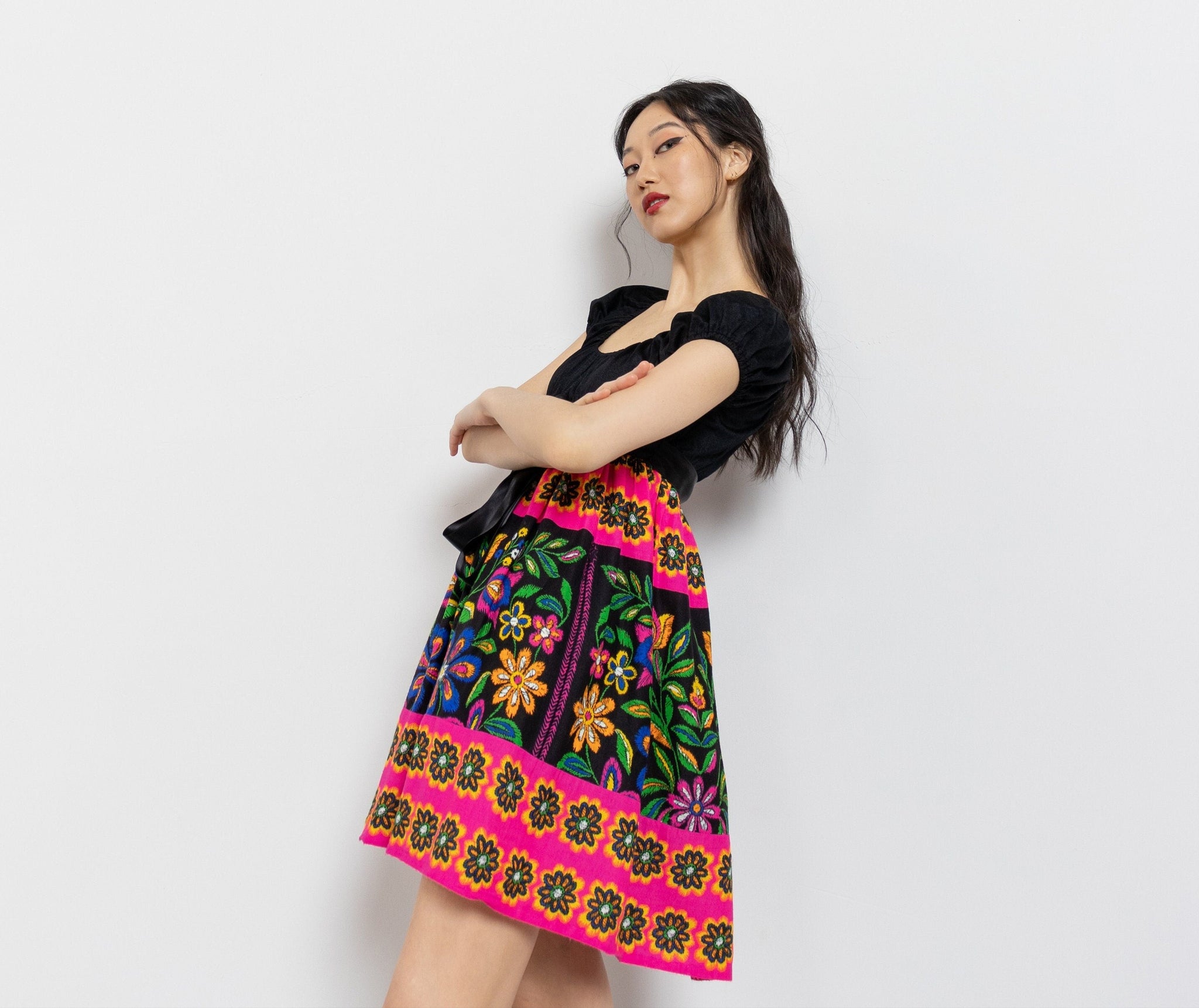 A woman posing in a vibrant mini dress with a colorful floral pattern on a black background, featuring a gathered bust and a pleated skirt.