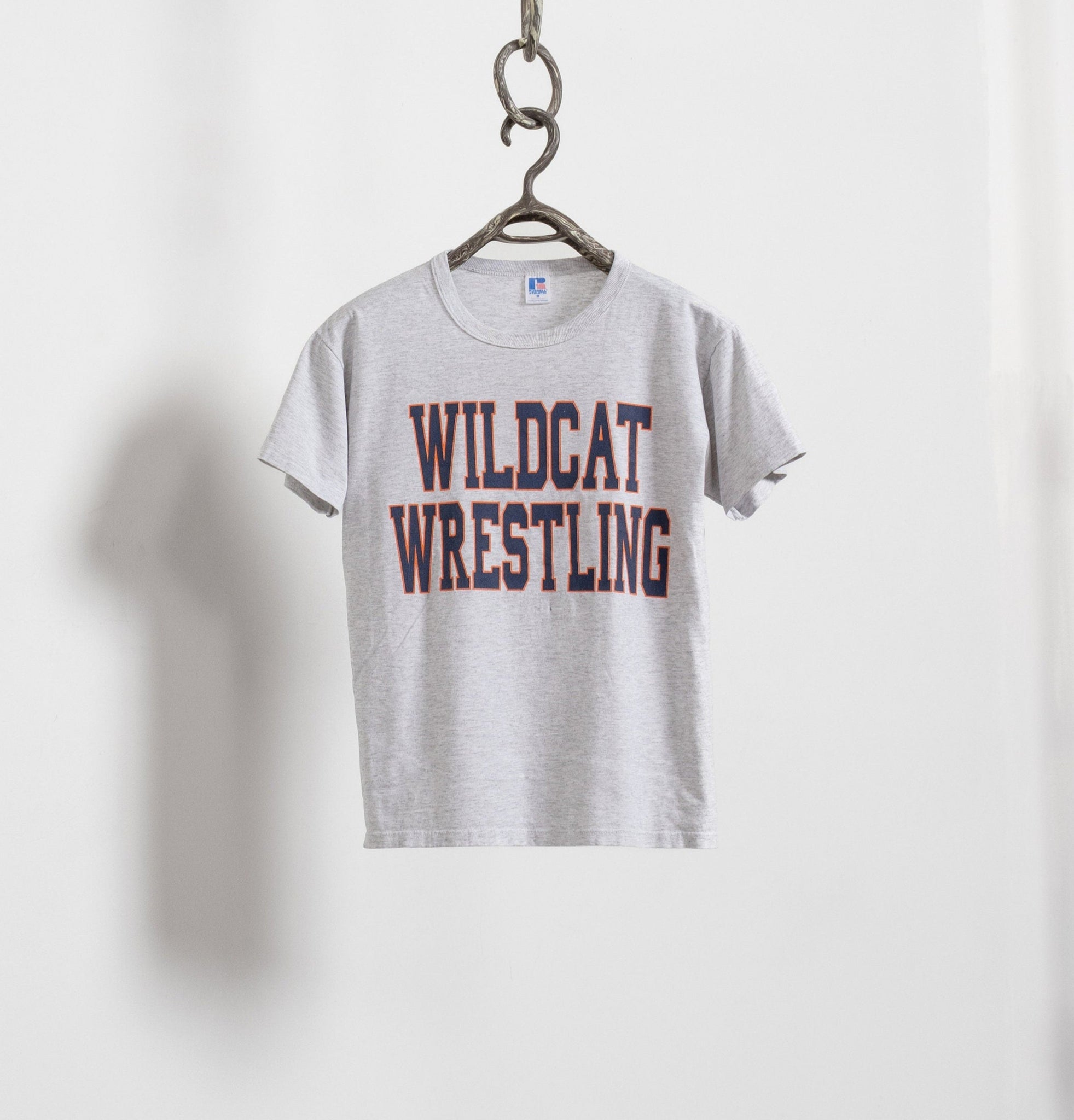 A heather grey Wildcat Wrestling t-shirt with orange text hanging from a chain.