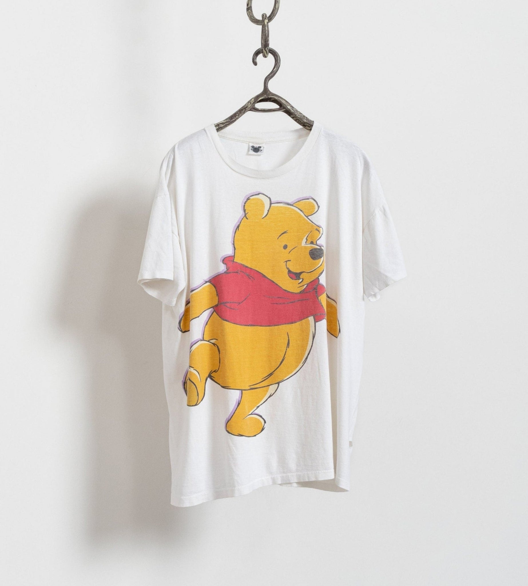 A white graphic t-shirt with a vintage illustration of Winnie the Pooh in a red shirt, hanging on a hanger against a white background.