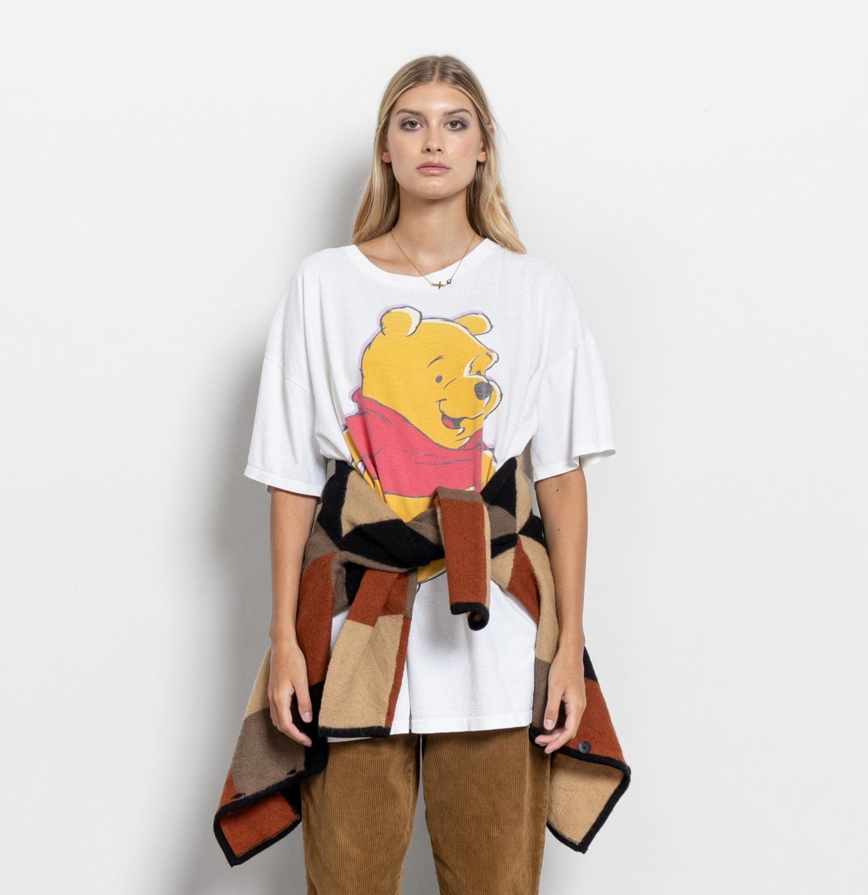 A person wearing a white graphic t-shirt with a vintage Winnie the Pooh design, paired with a multi-colored patchwork kimono and brown pants.