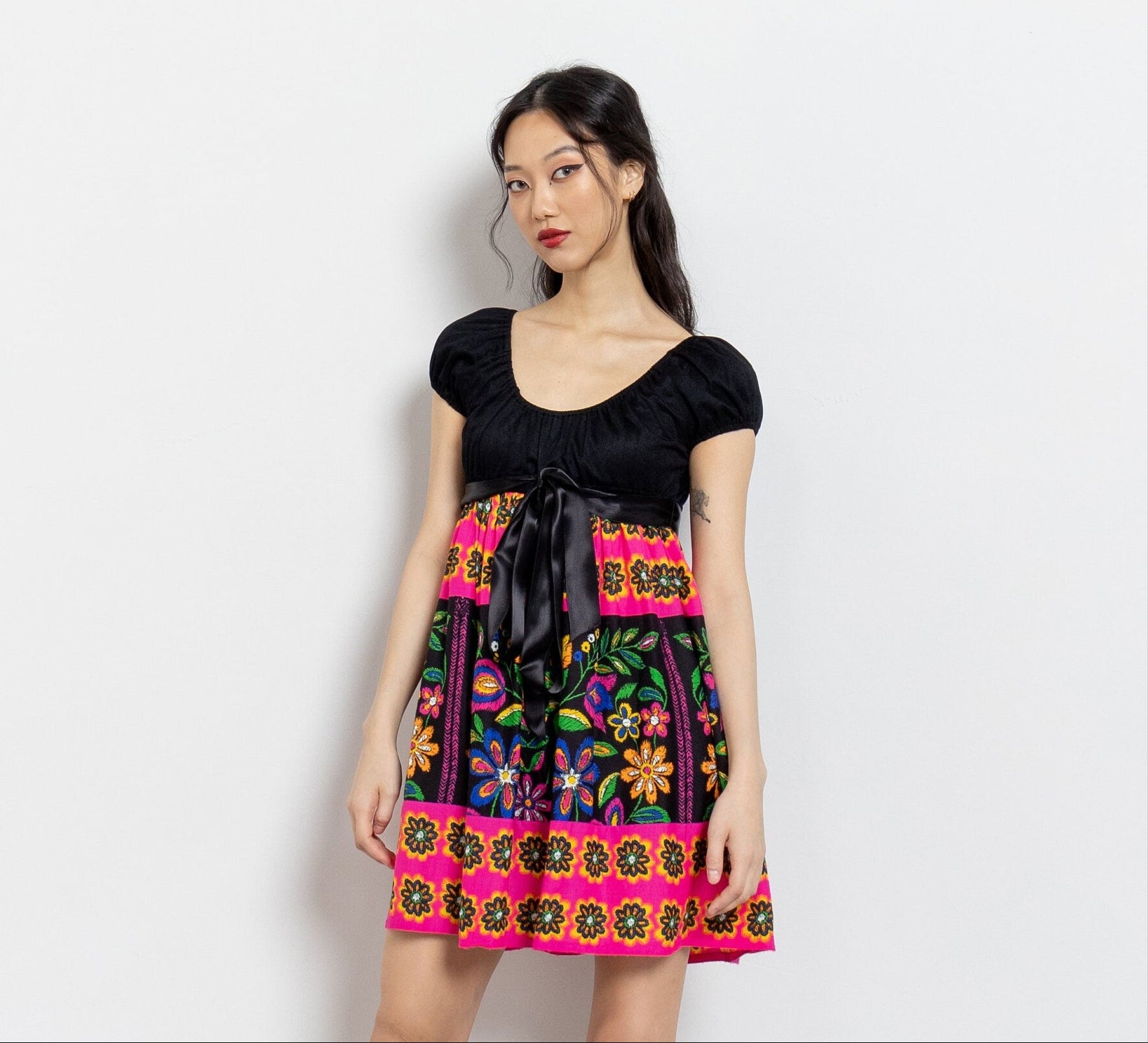 A vintage 70's black and bright colors floral bardot mini dress with a contrasting black bodice, pleated skirt, and elastic sleeve hems. The dress features a colorful floral pattern against a black background and ties up in the back with a ribbon below the bust-line.
